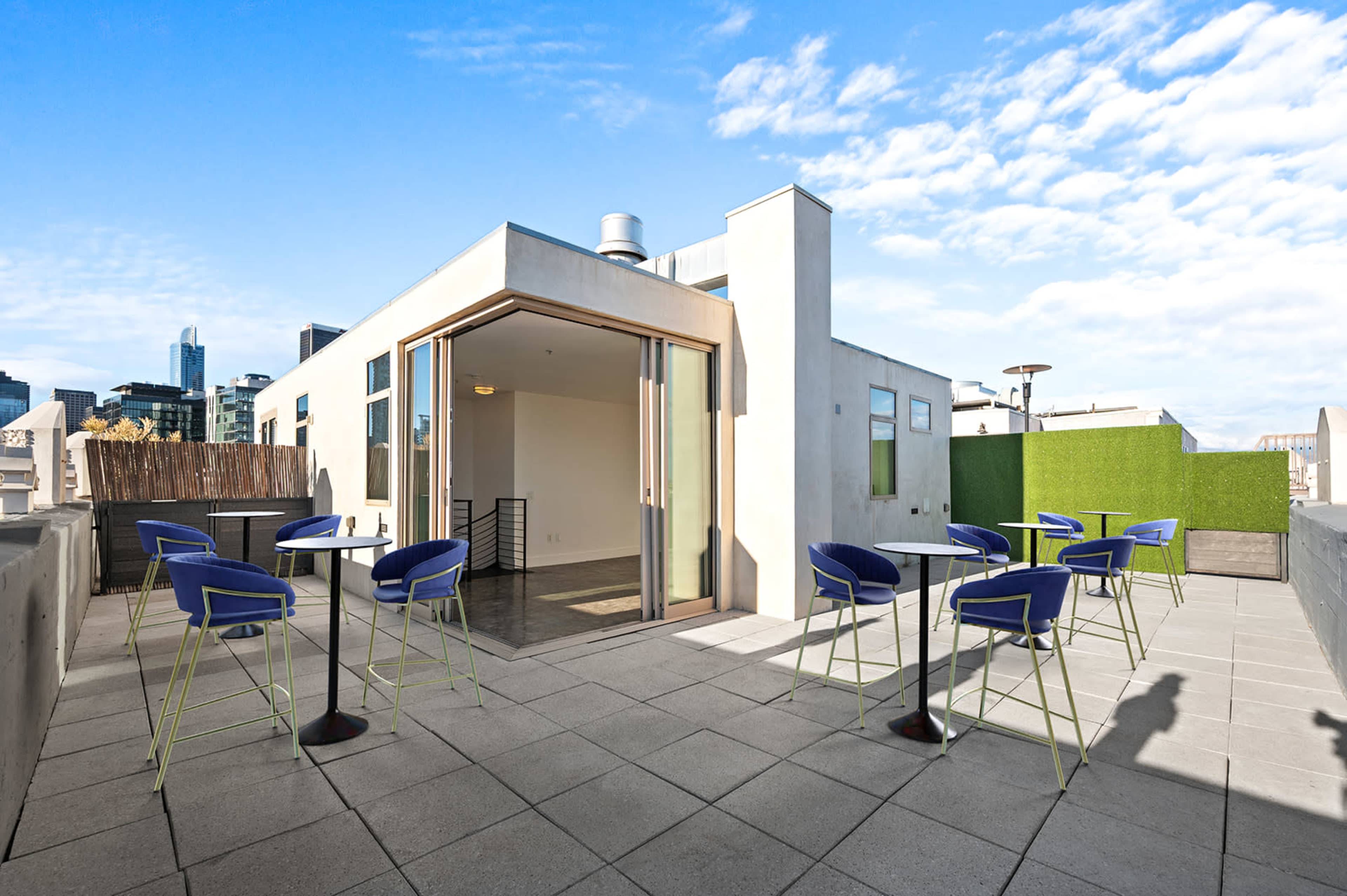 The image shows a rooftop terrace featuring several high tables with blue chairs, surrounded by modern buildings under a clear sky.