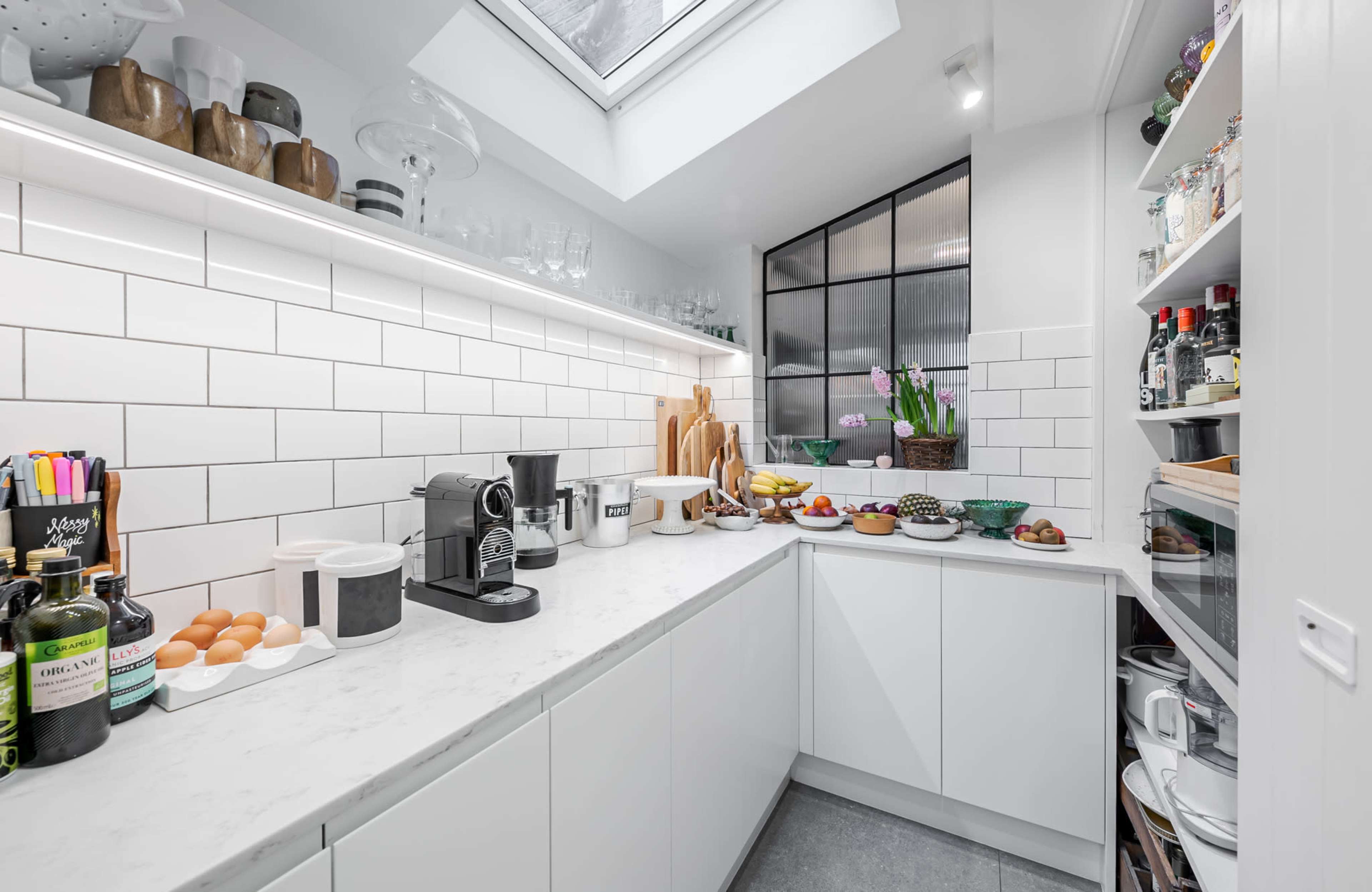 A compact kitchen features white cabinetry, a marble countertop, and a skylight, with a variety of food items and kitchen appliances arranged on the surfaces.
