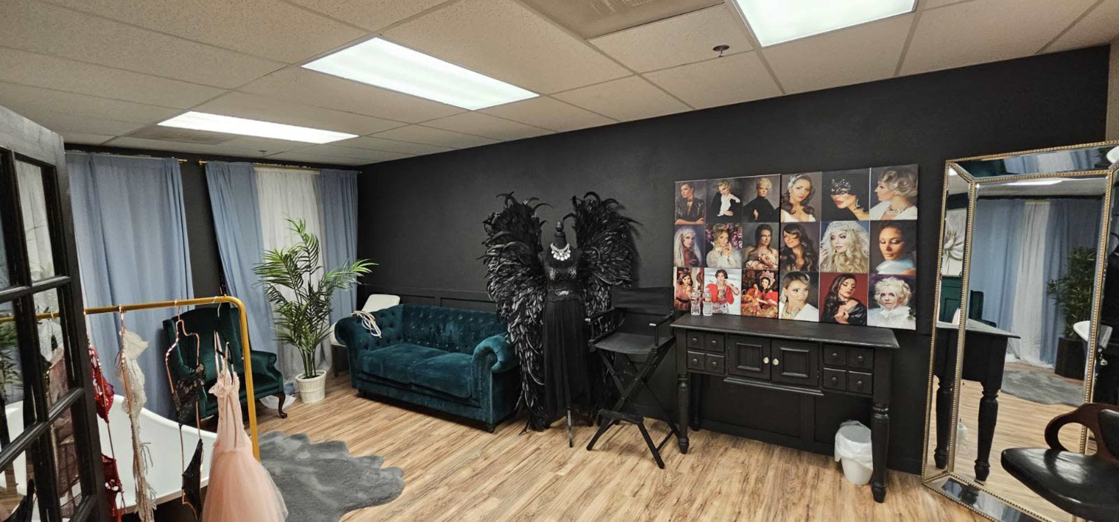 The photo shows a stylish studio space featuring a teal couch, a makeup station, a black wall adorned with portraits, and an ornate black costume with feathers.