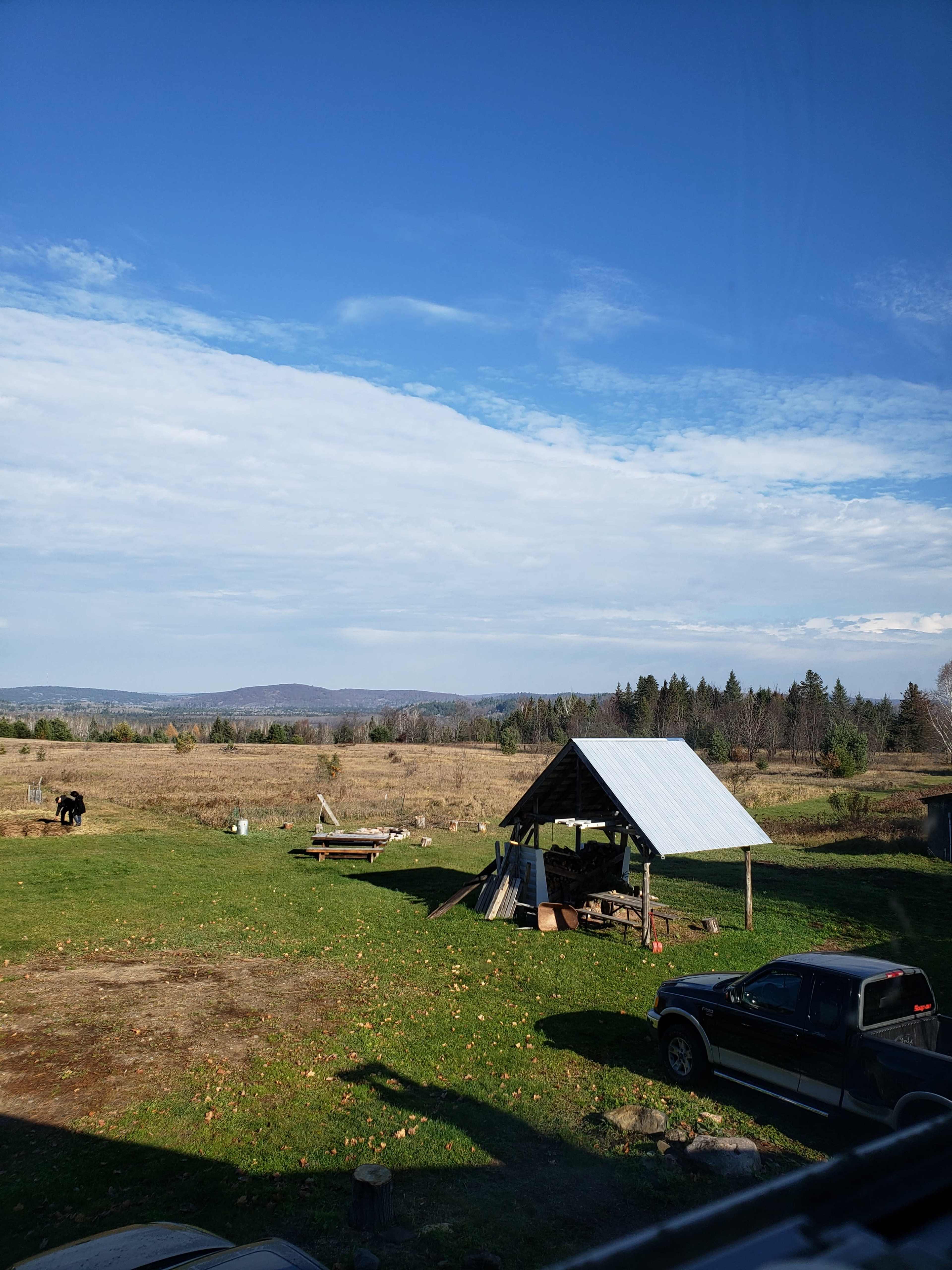 A cleared rural landscape with a wooden shelter, a parked truck, and distant hills under a partly cloudy sky.