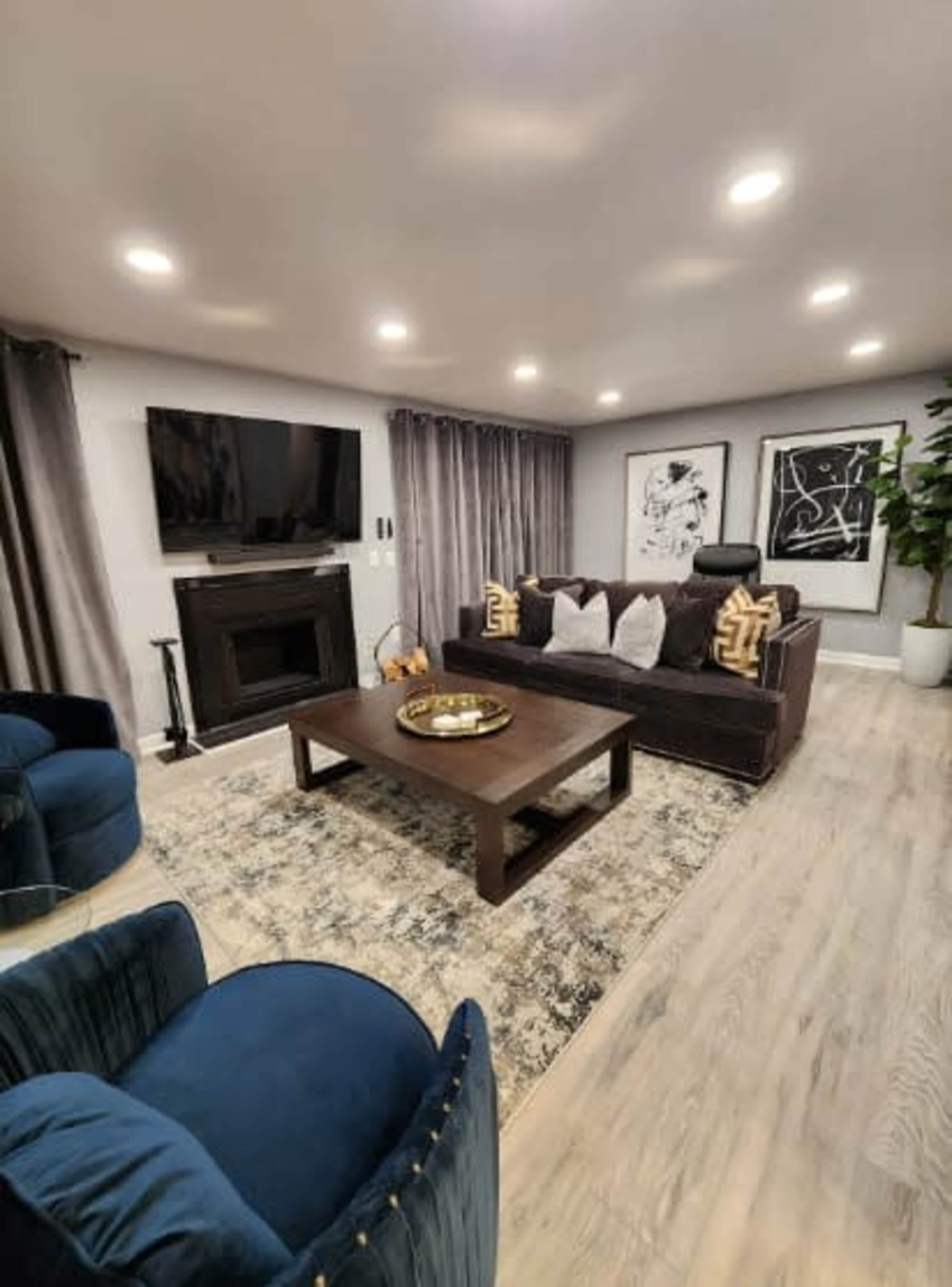 The living room features a dark sofa with decorative pillows, a wooden coffee table, a TV mounted on the wall, and two blue armchairs, all arranged around a patterned area rug.