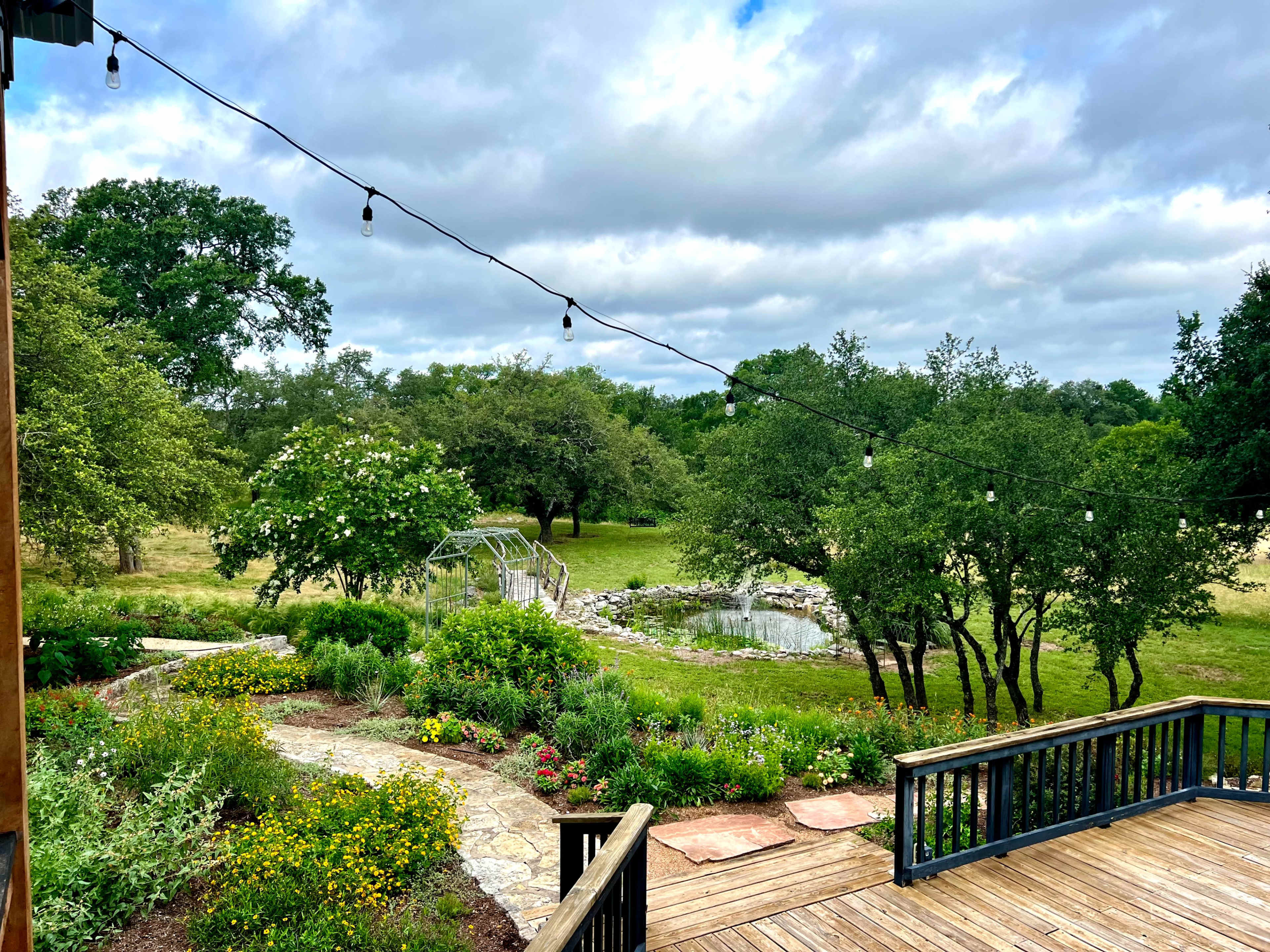 A wooden deck overlooks a landscaped garden with winding paths, a pond, and string lights overhead.