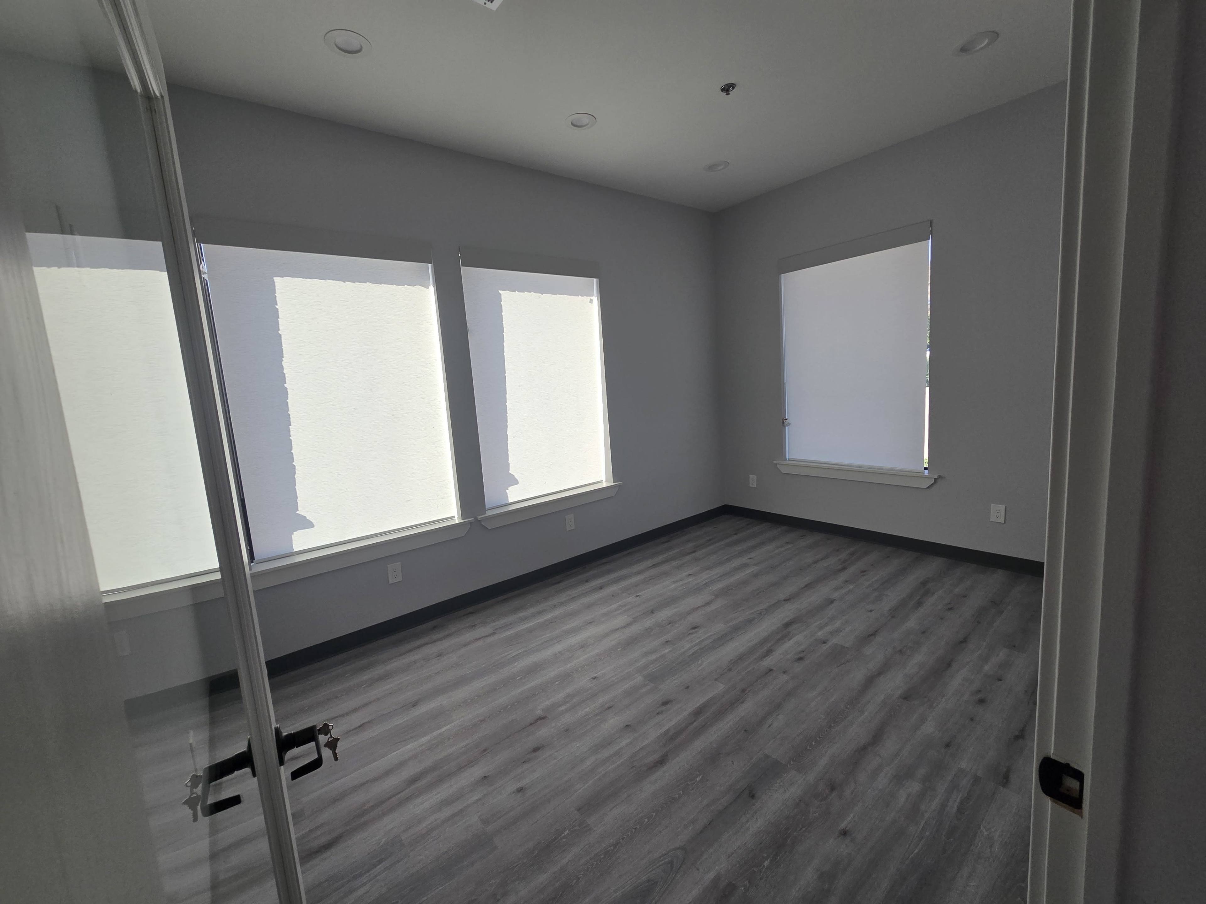The image shows an empty room with gray walls and hardwood flooring, featuring three windows covered by shades.
