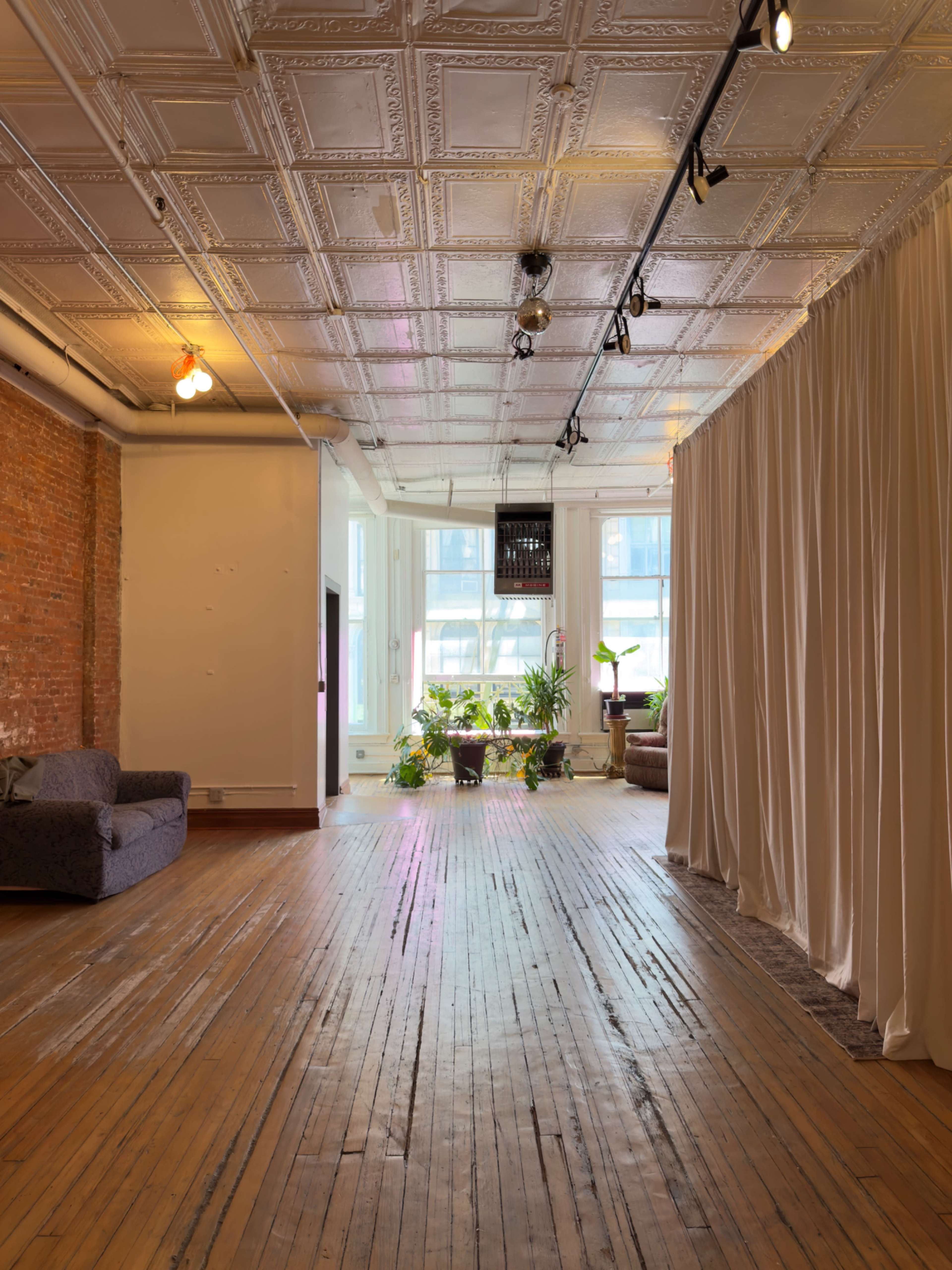 A spacious room features wooden floors, exposed brick walls, large windows with natural light, and a sectioned area with curtains.