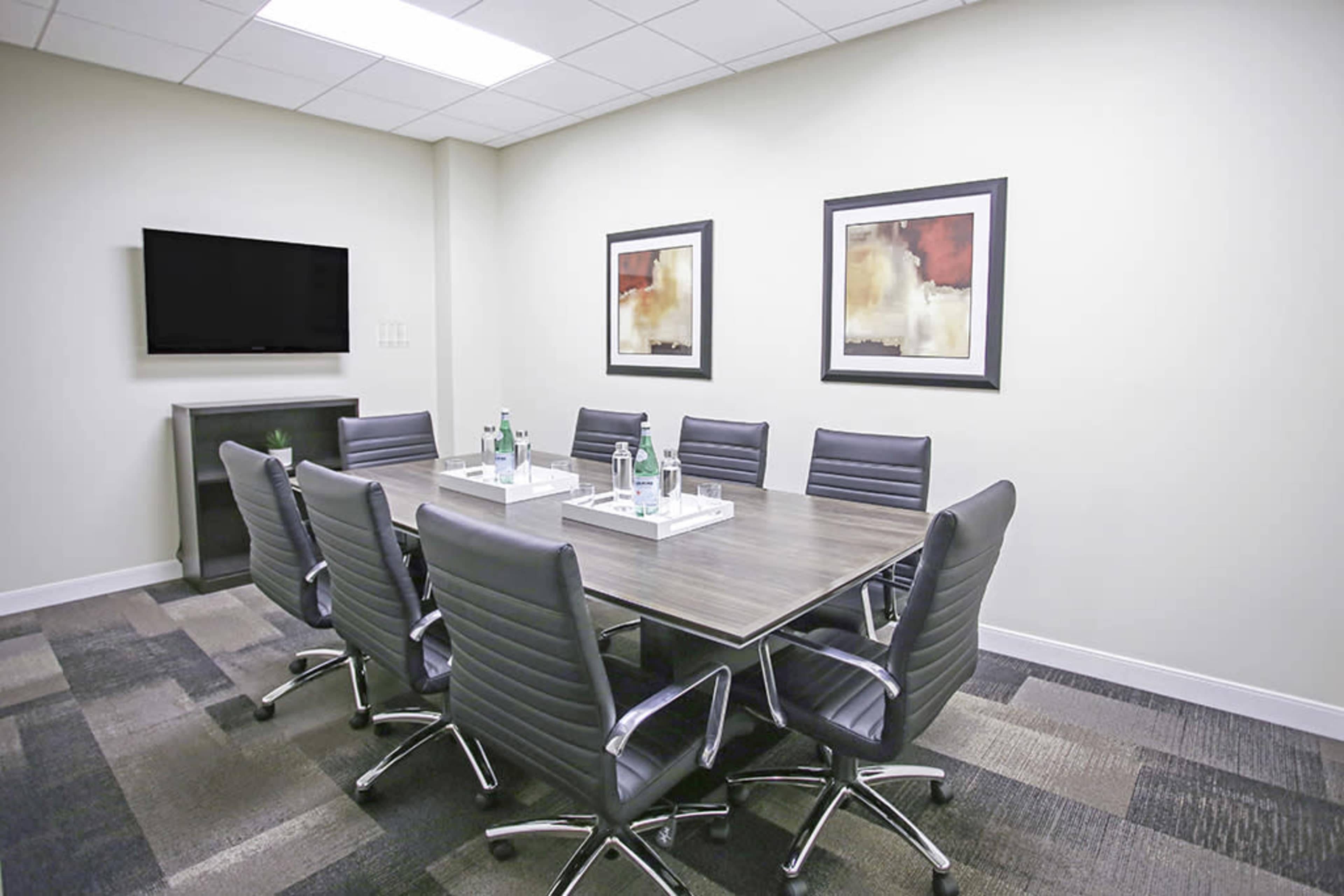 A modern conference room features a large table surrounded by eight ergonomic chairs, with two framed artworks and a television on the wall.