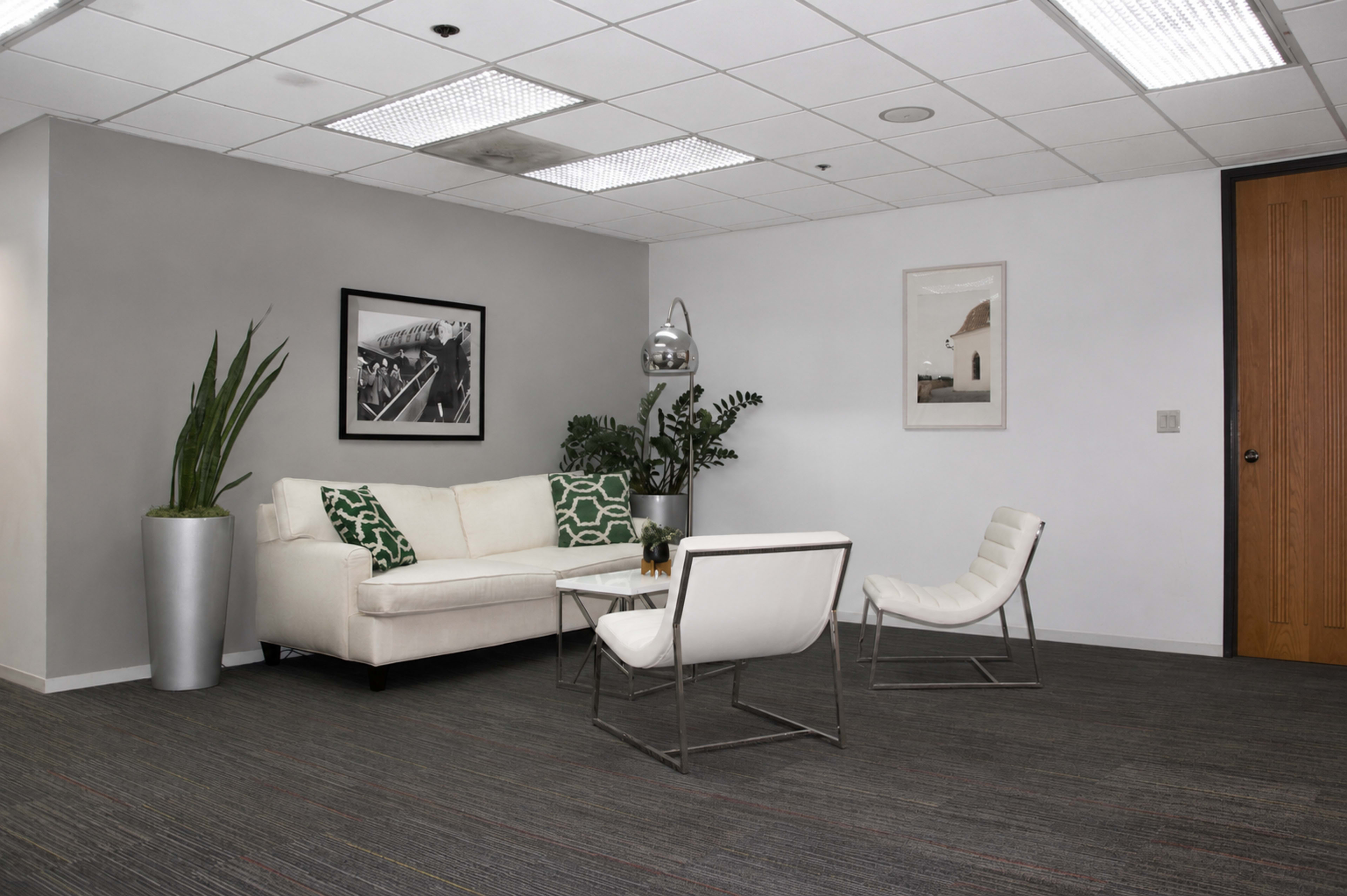 The image shows a modern office lounge area with a white couch, two white chairs, a glass coffee table, and decorative plants in a sleek environment.
