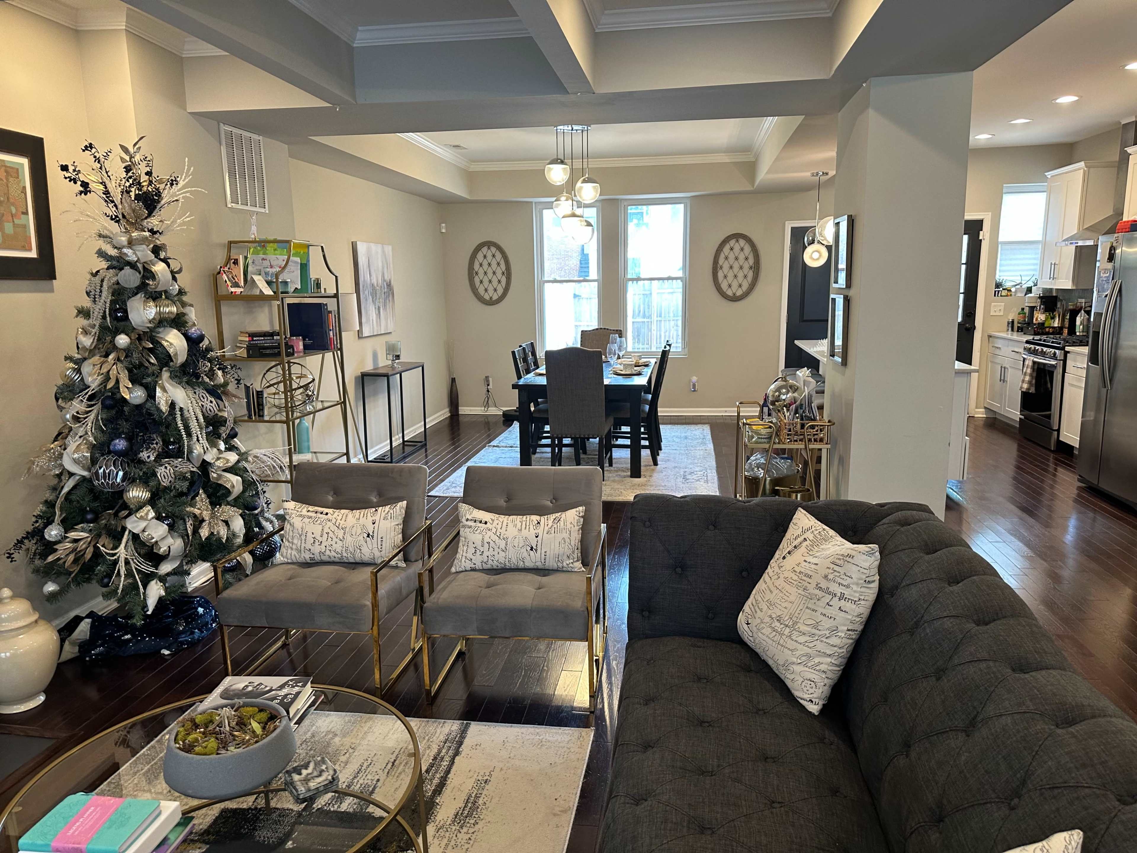 A living room with a Christmas tree, two gray sofas, and a dining area in the background featuring a dining table and chairs.