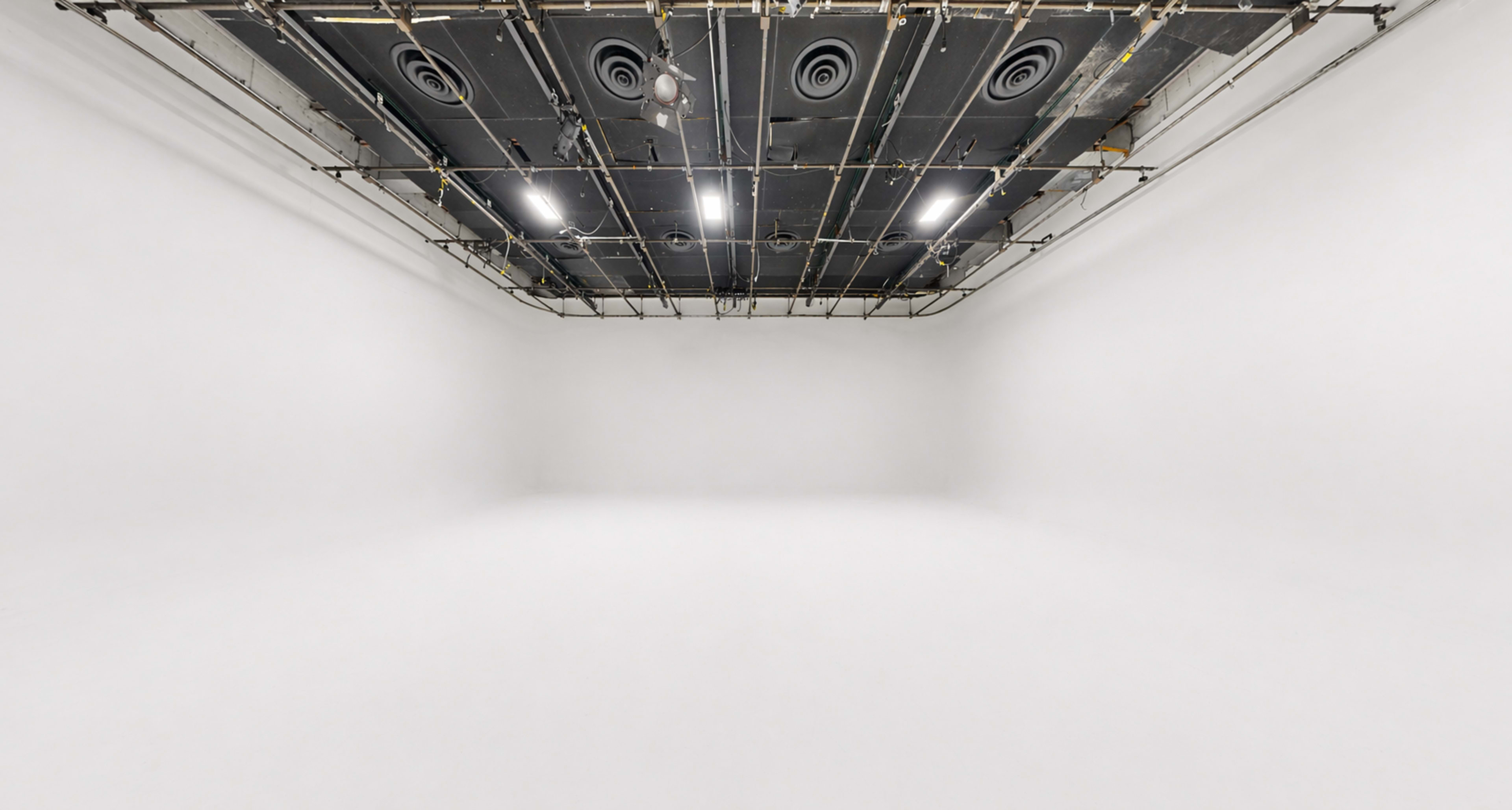 The image shows a spacious, empty white room with a ceiling featuring exposed lighting and ventilation elements.