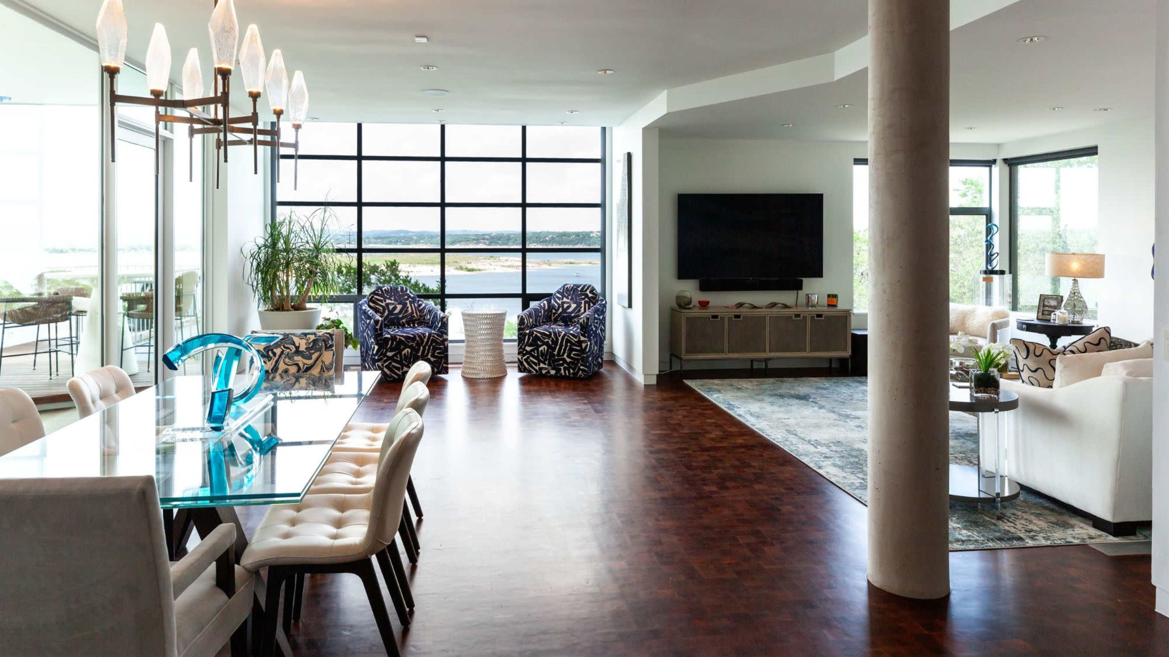 The image shows a spacious, modern living area with large windows overlooking a body of water, featuring a dining table, lounge chairs, and a television.