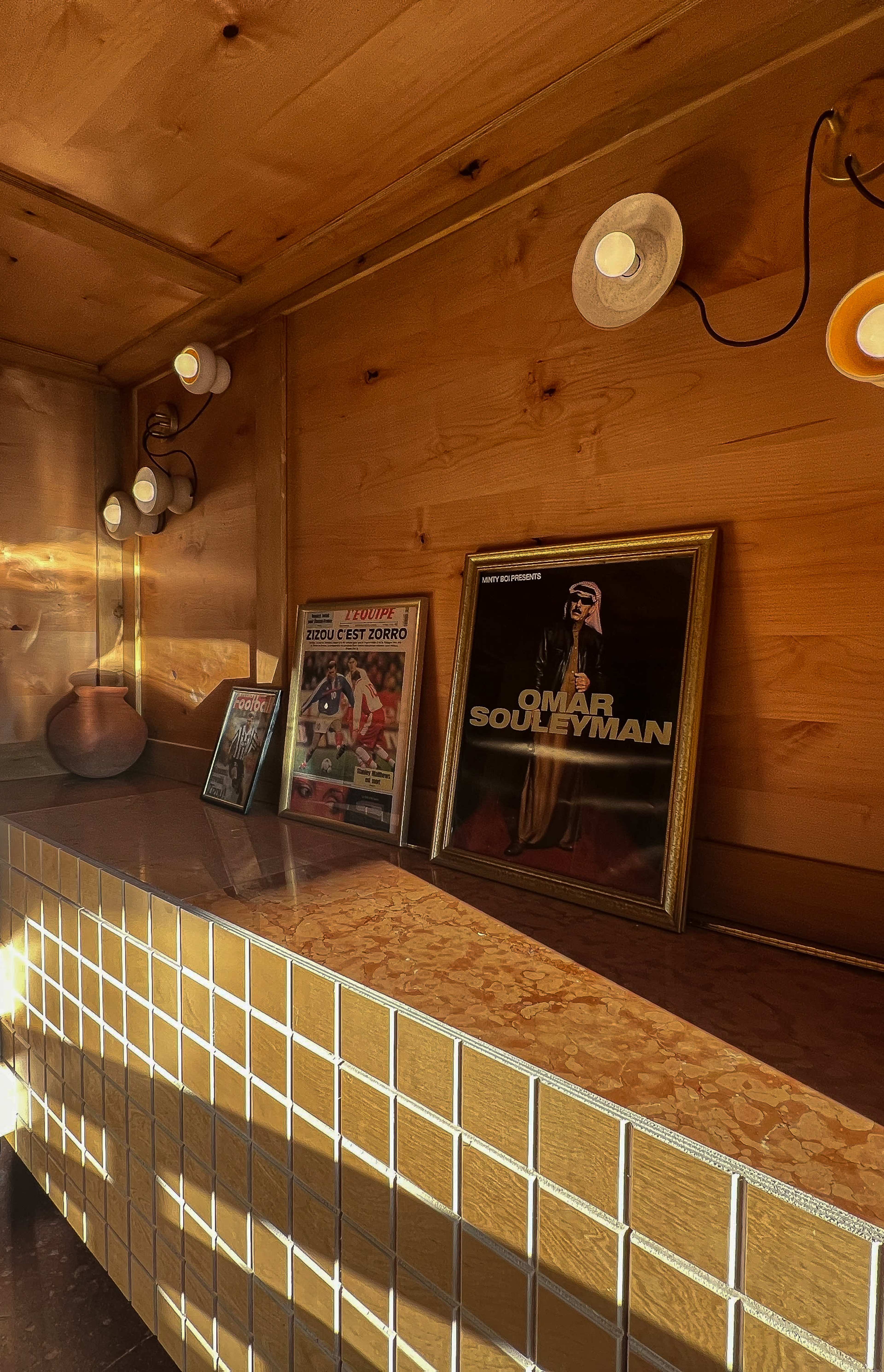 The image features a wooden interior with a low table displaying framed records and magazines, alongside a ceramic pot and wall sconces.