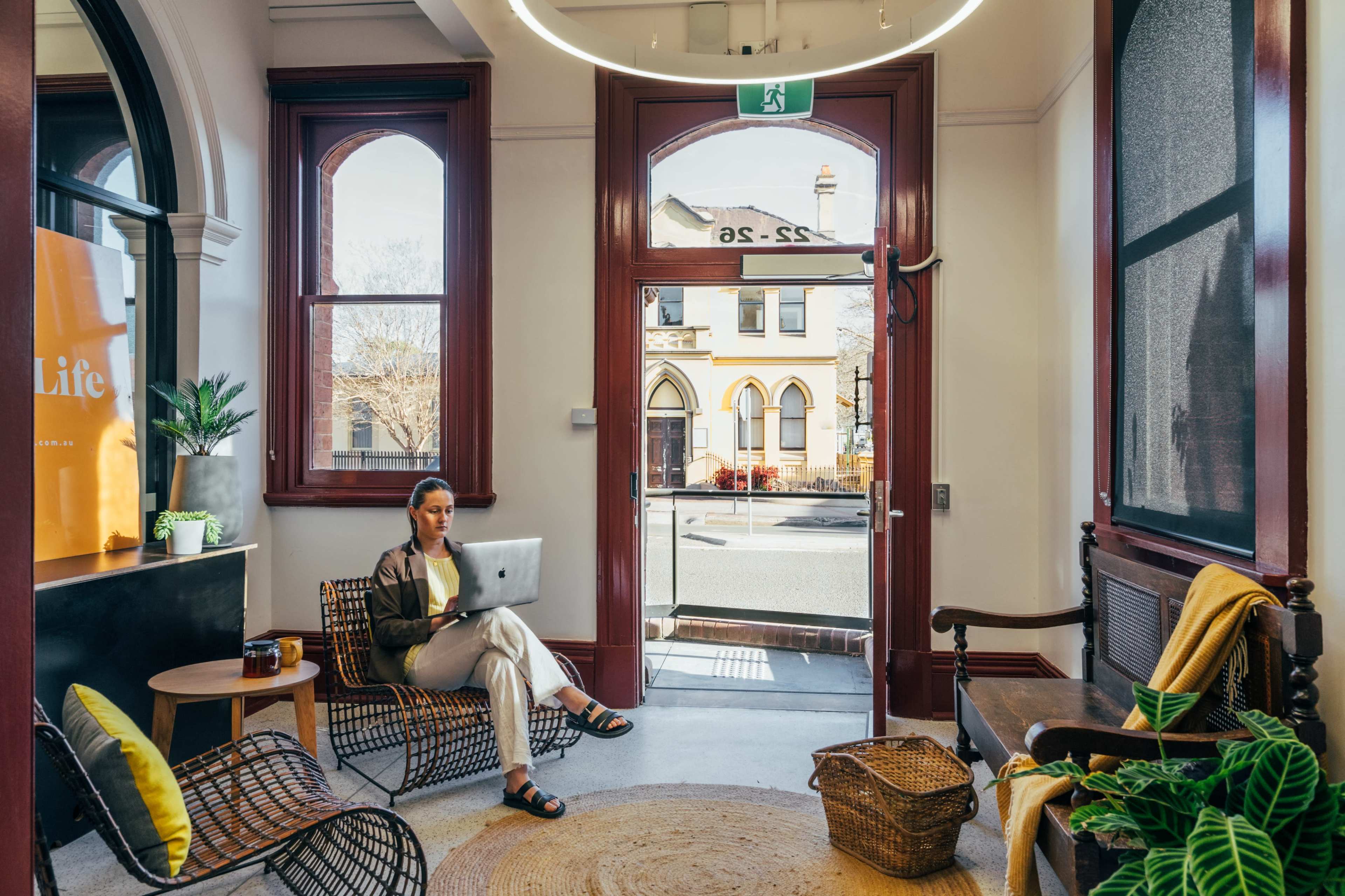 A woman sits on a chair with a laptop in a room featuring large windows and a view of a street outside.