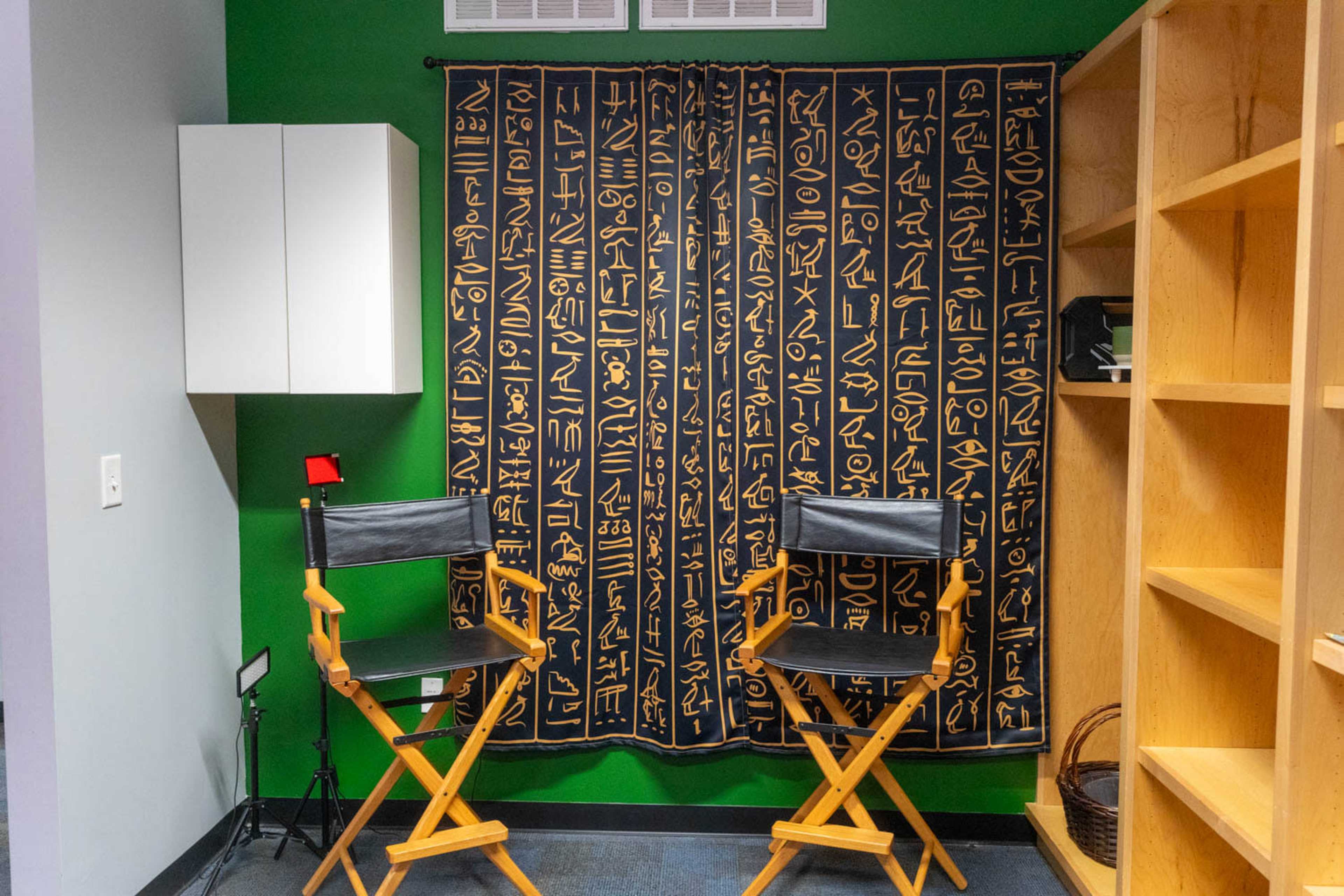 A room with two black director's chairs facing a curtain adorned with hieroglyphic designs, set against a green wall and light wooden shelving.