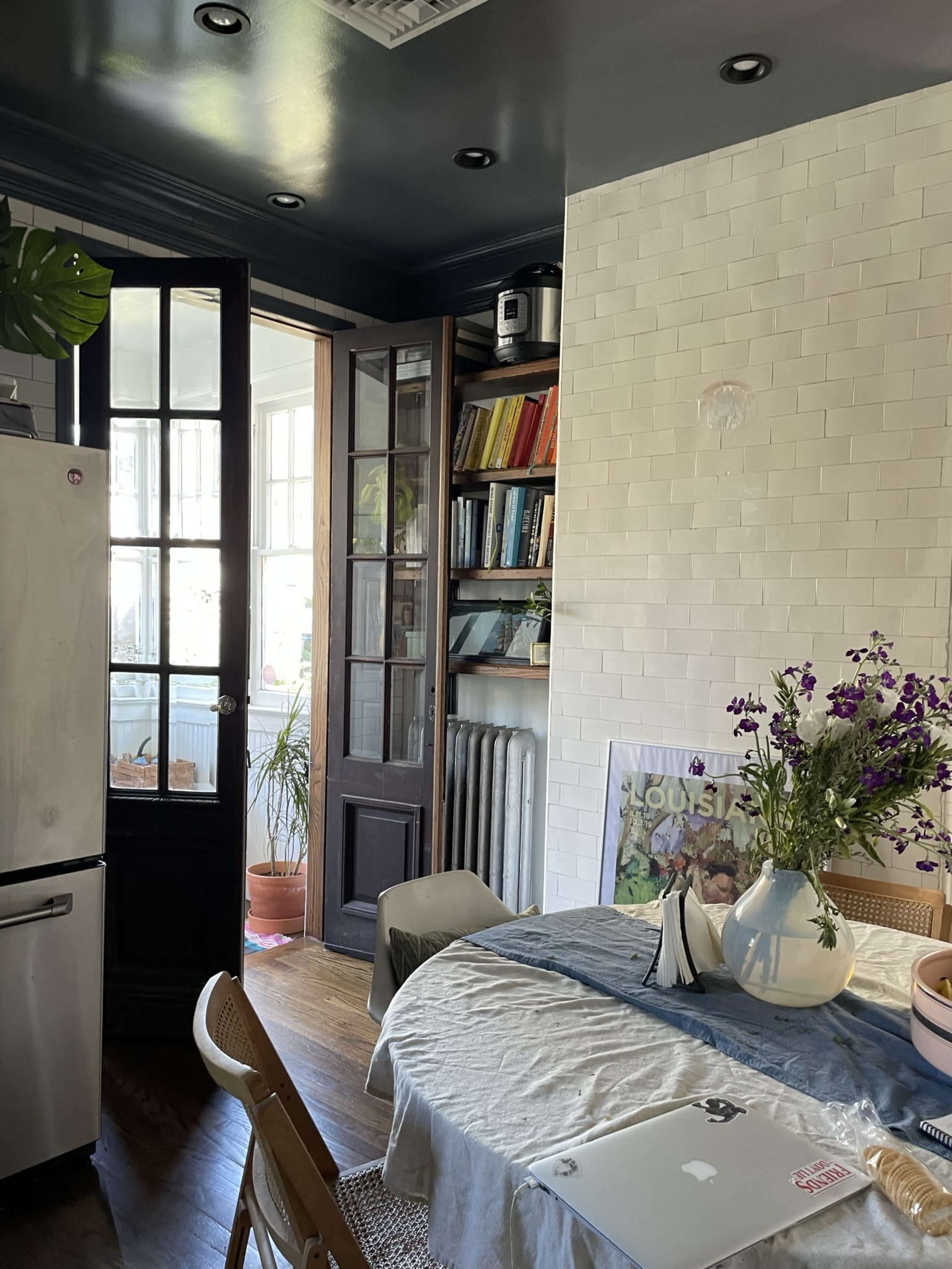 A kitchen features a dining table with a vase of flowers, a computer, and shelves filled with books, while a doorway leads to a bright room beyond.