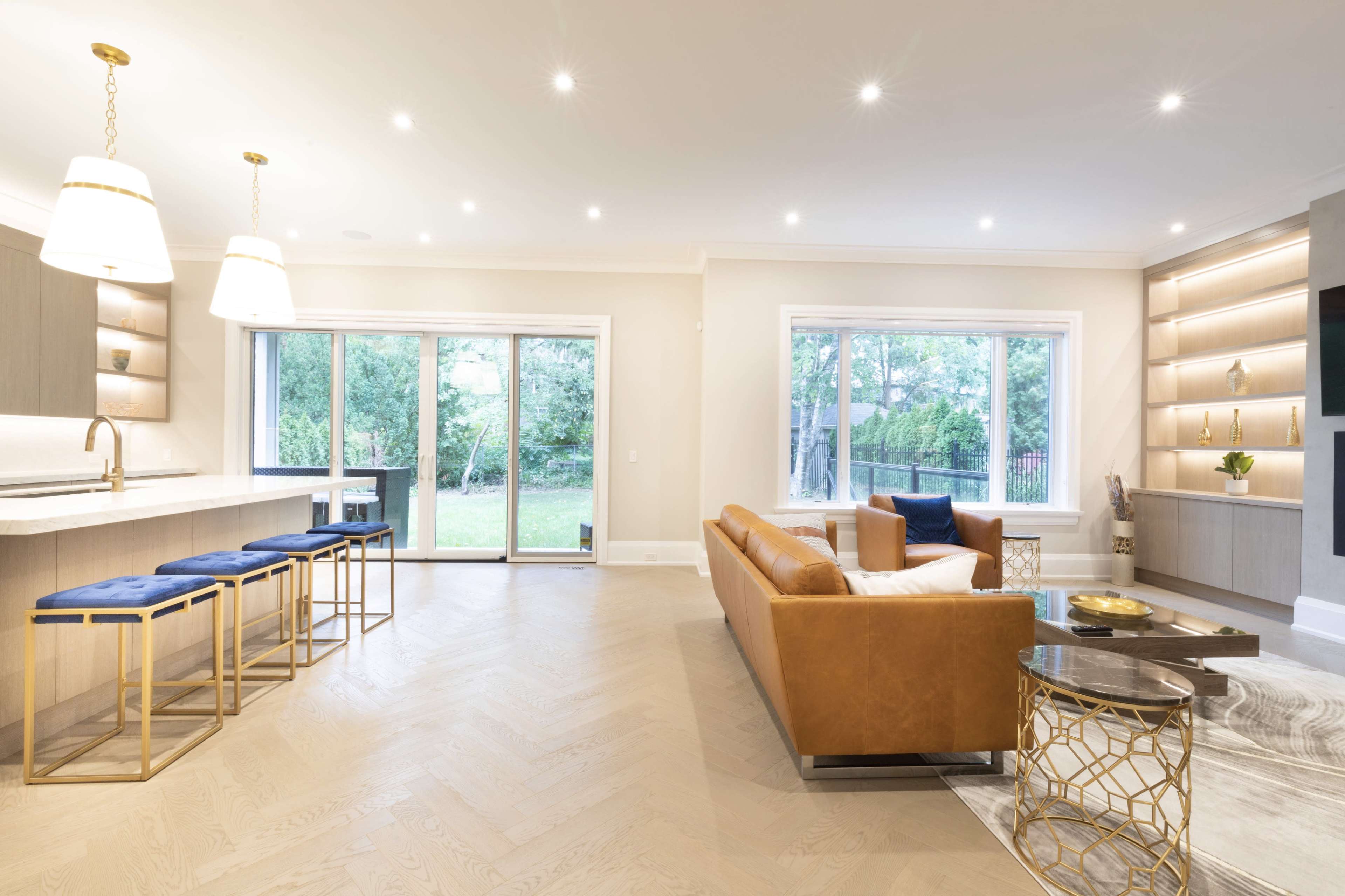 A modern living room with a brown leather sofa, a glass coffee table, and a kitchen area featuring bar stools and a large window overlooking a garden.
