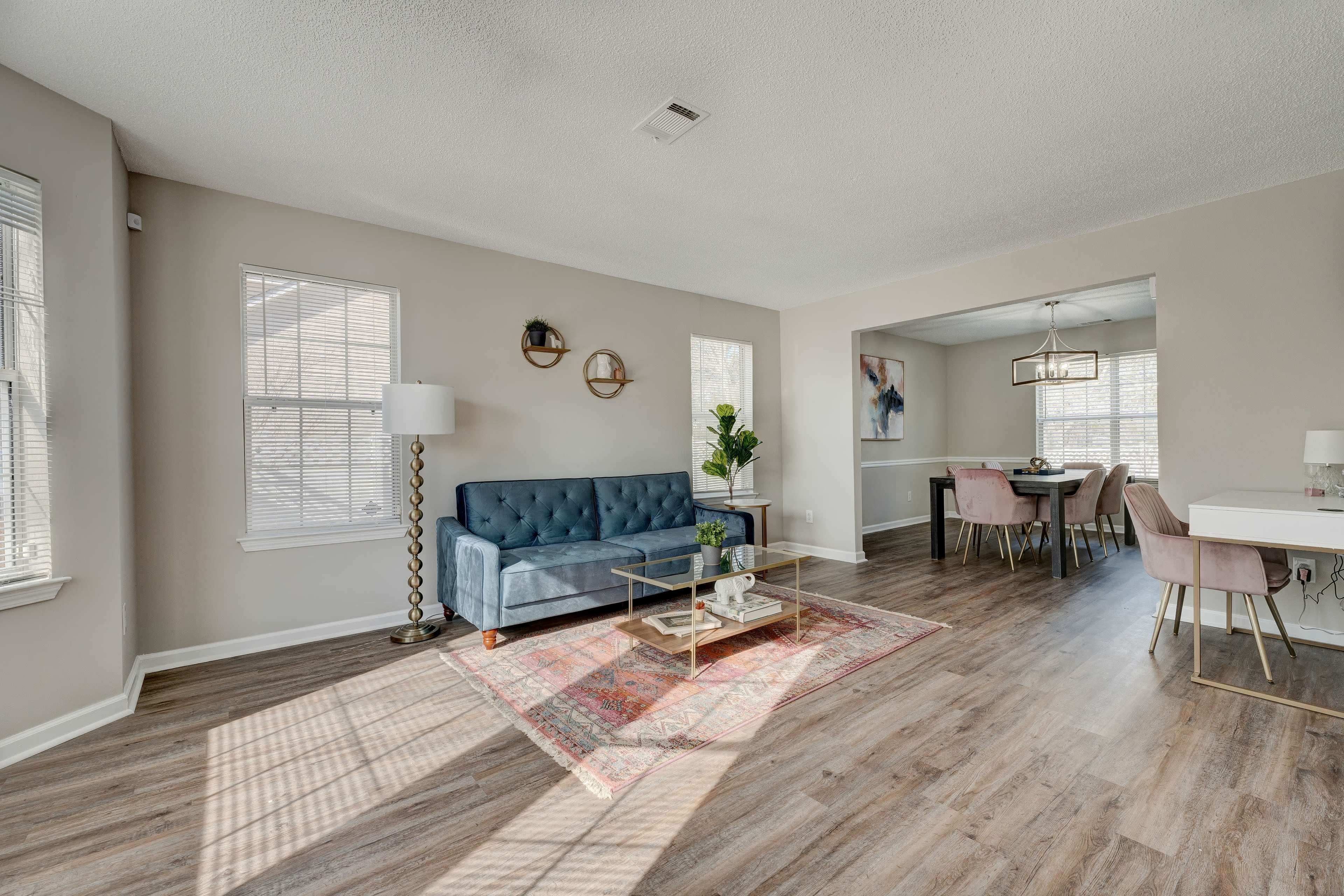 A well-lit living room with a blue sofa, a coffee table on a patterned rug, and an adjoining dining area with a table and chairs.