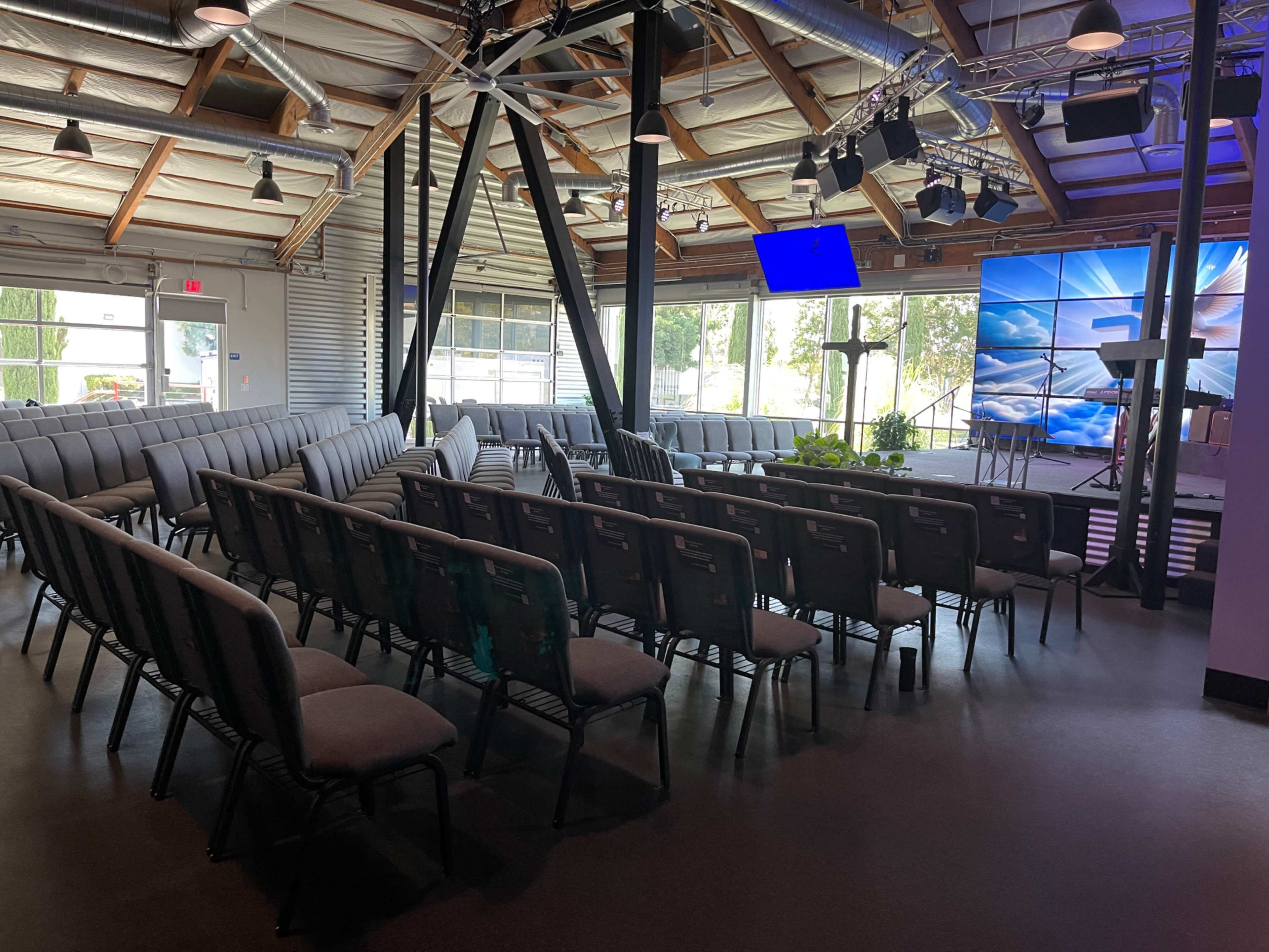 The image shows a modern church interior with rows of gray chairs facing a stage that features a cross and a large projection screen.