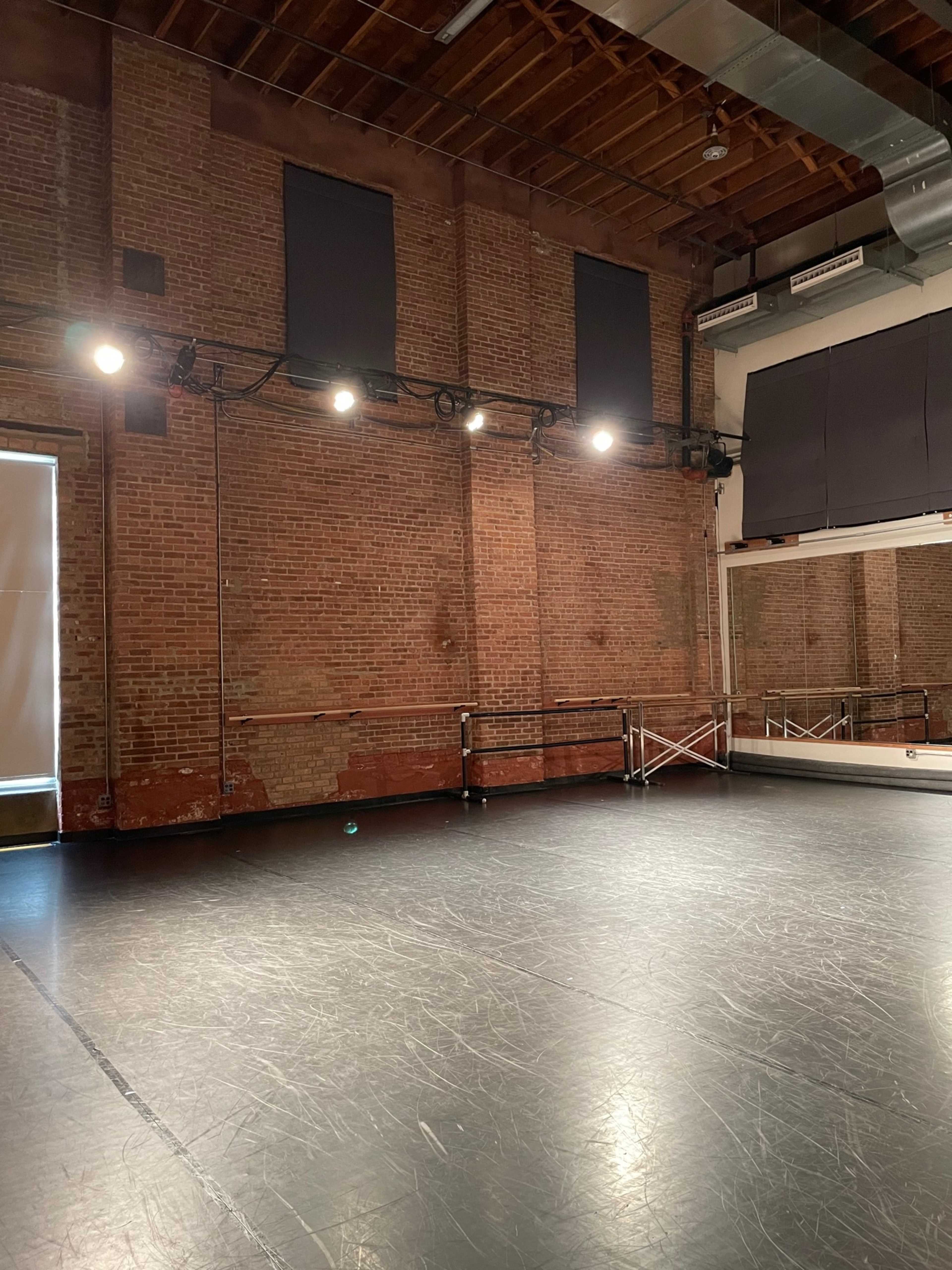 The image shows a spacious dance studio with exposed brick walls, overhead lights, and a mirrored wall.