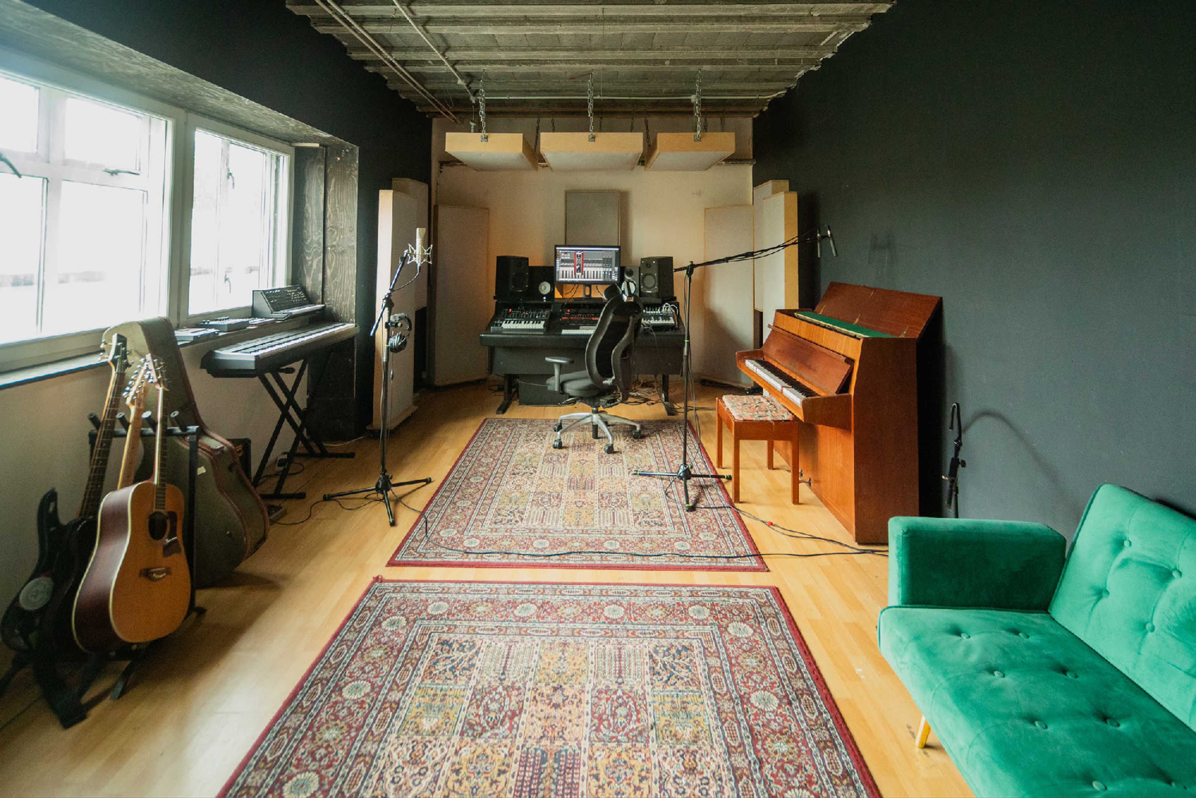 The image shows a music recording studio with various instruments, including keyboards, guitars, and a piano, arranged around a central workspace with a computer setup and microphones.