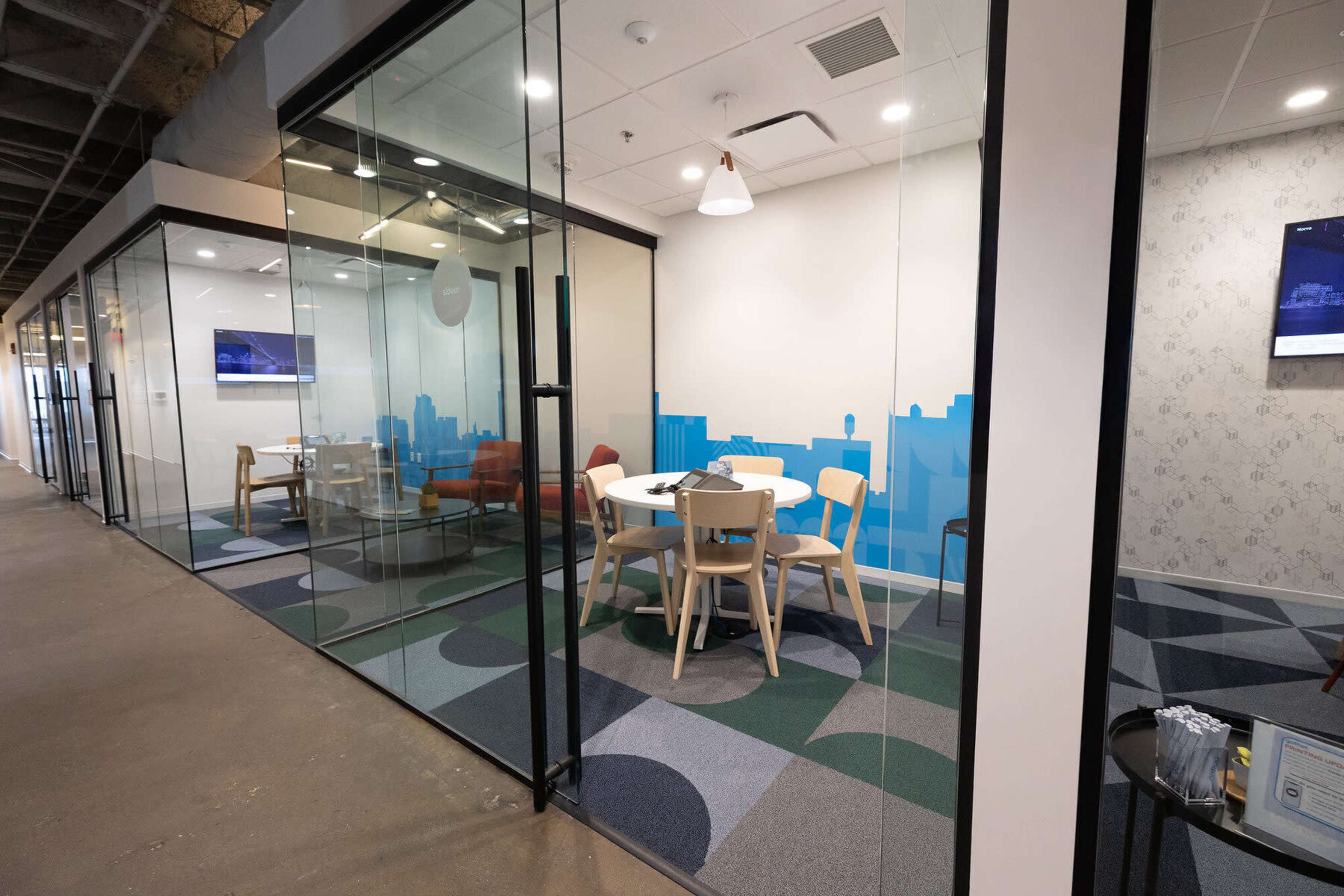 The image shows a modern office space with glass-walled meeting rooms, a circular table with chairs, and colorful patterned flooring.