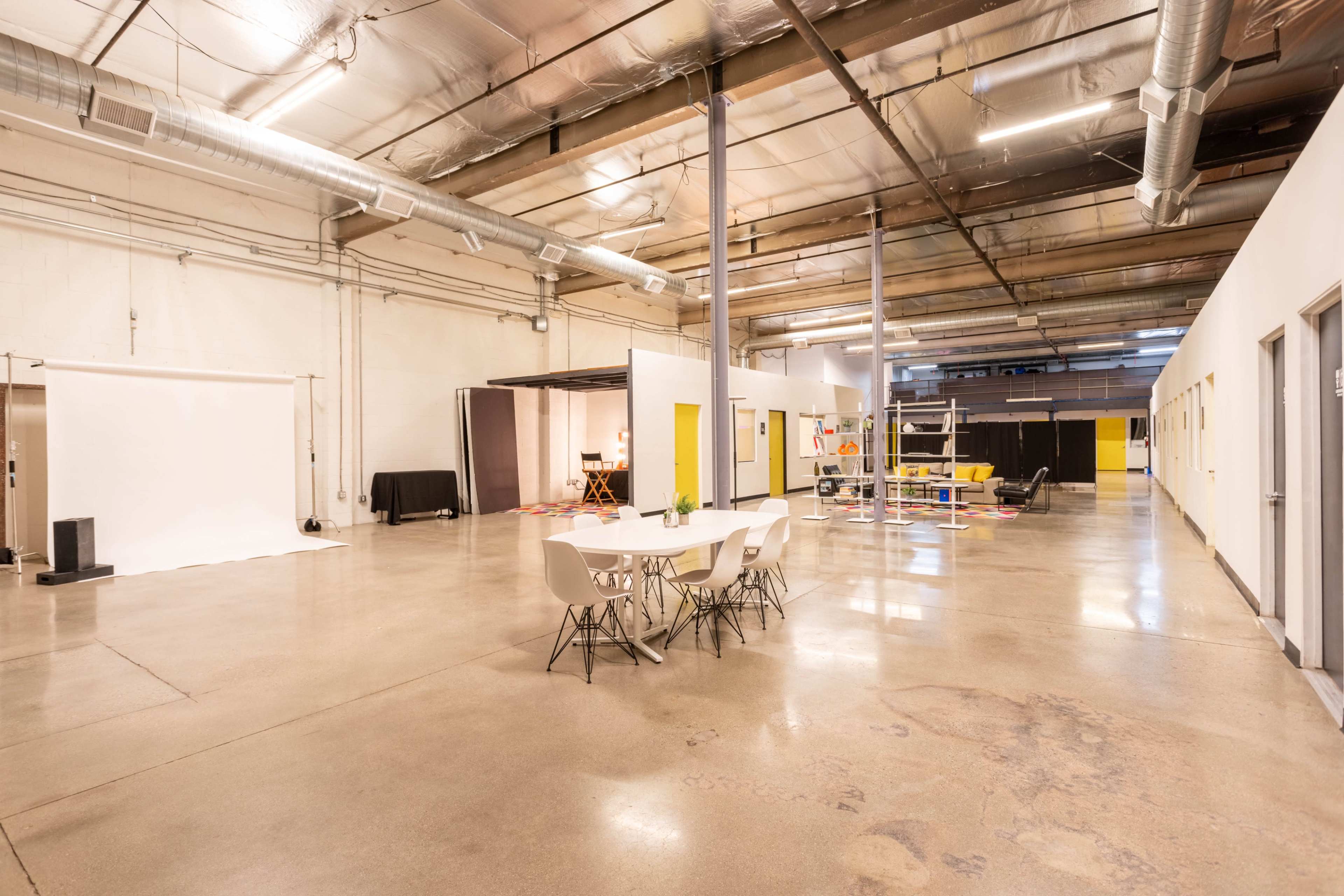 A spacious, well-lit industrial interior featuring a dining table with chairs, empty areas, and various rooms with yellow doors.