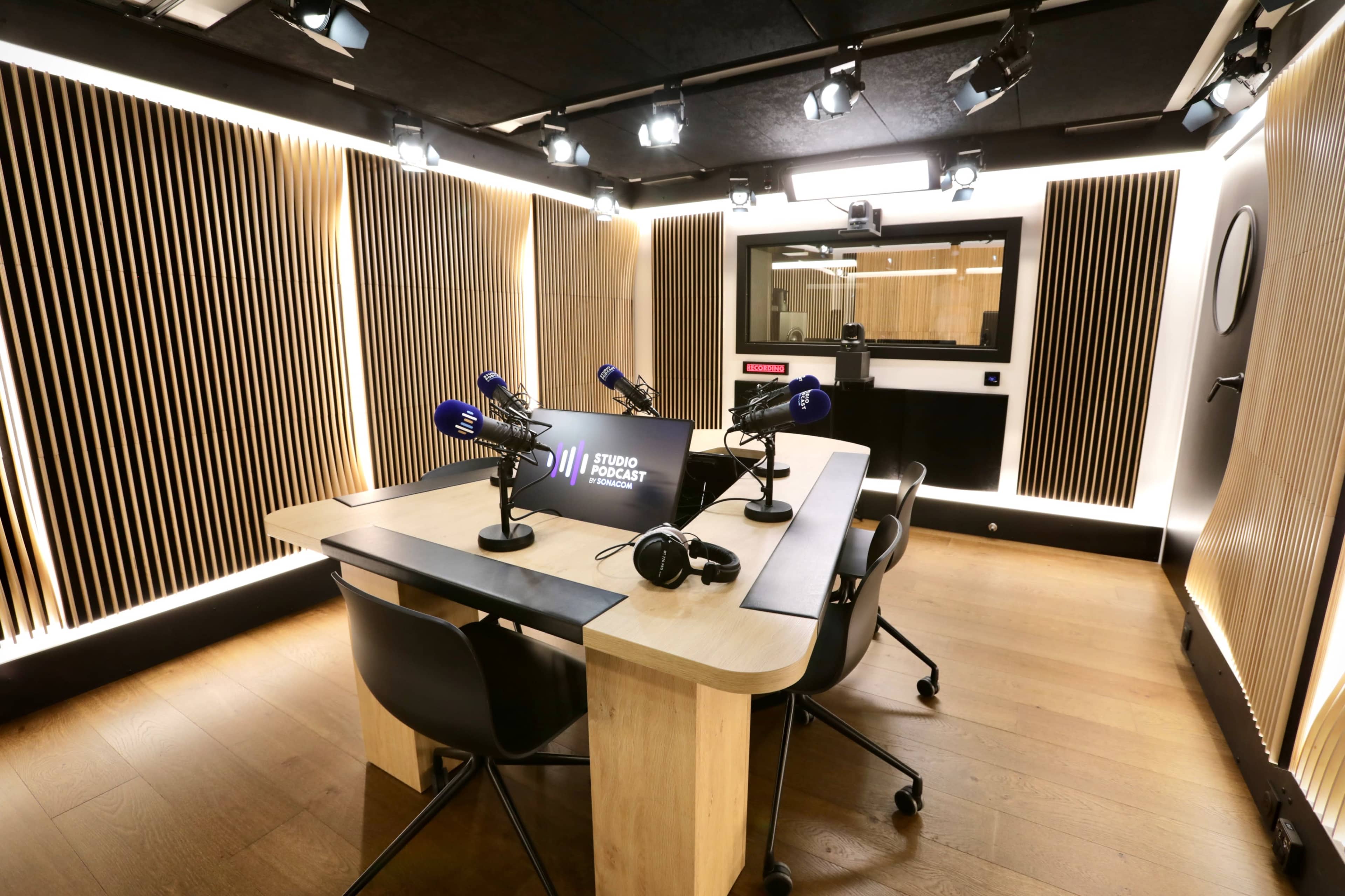 A modern podcast studio featuring a wooden table, multiple microphones, and adjustable lighting.