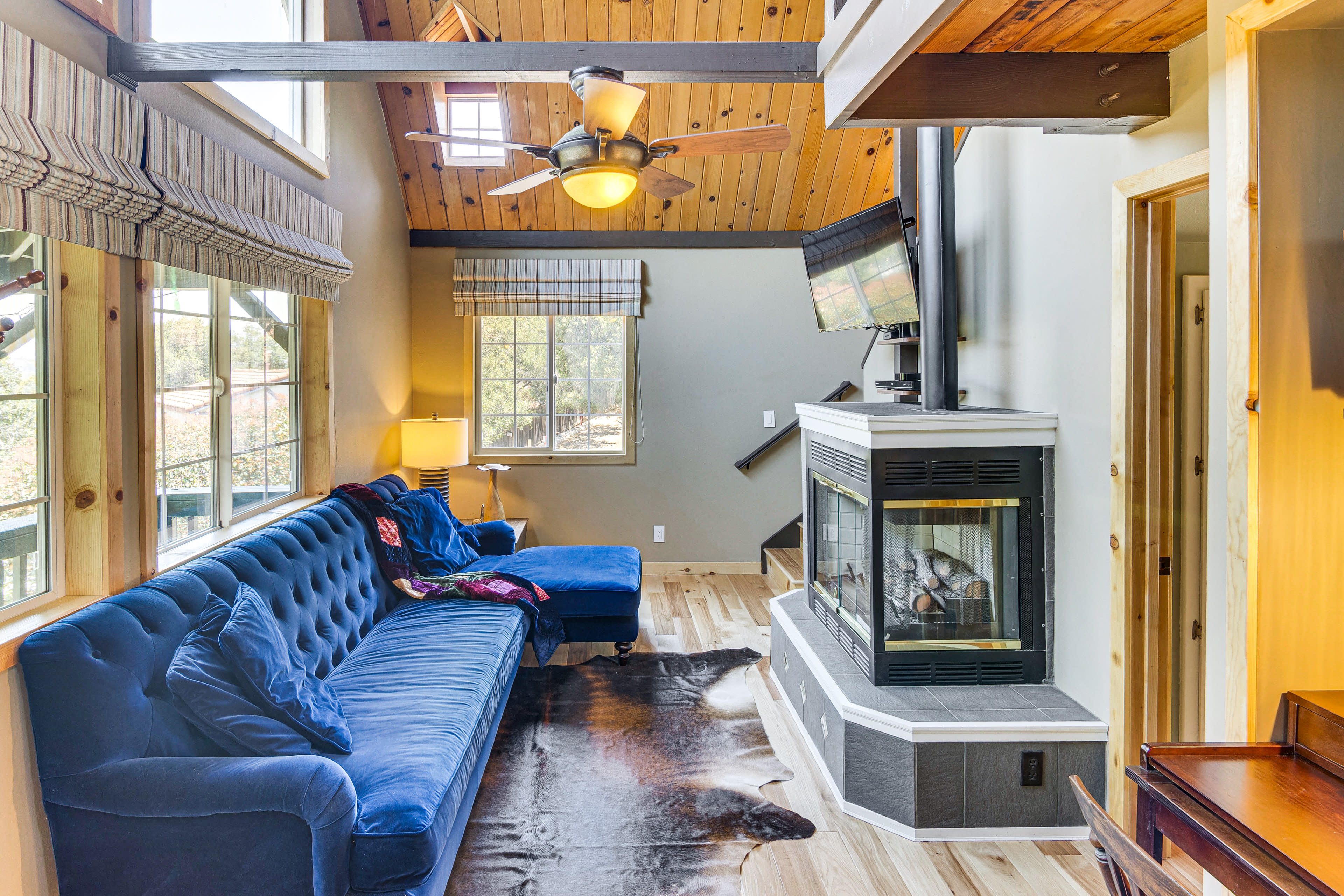 The image shows a cozy living room featuring a blue sofa, a chaise lounge, a fireplace, and large windows allowing natural light to fill the space.