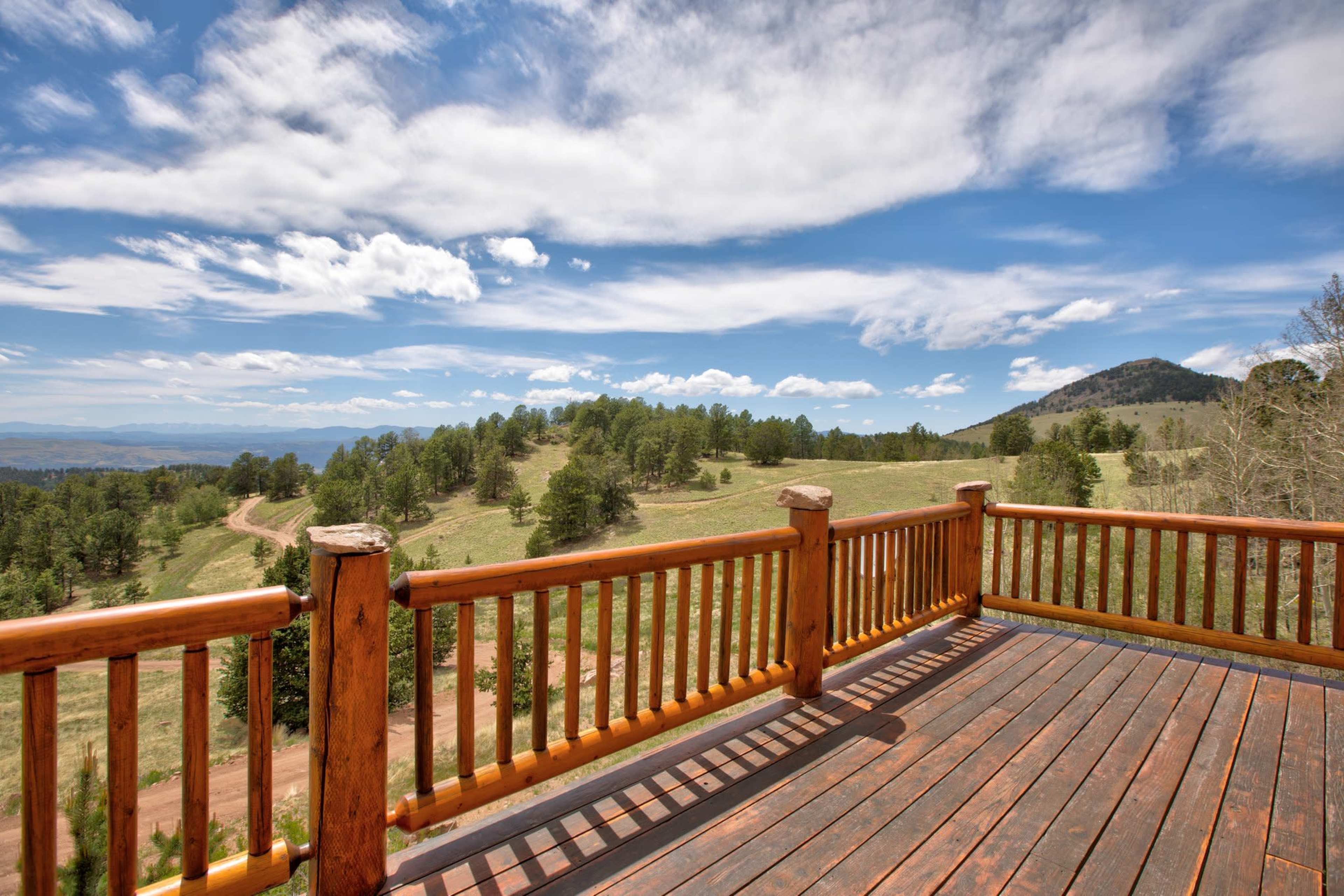 A wooden deck overlooks a green landscape with a winding dirt road and distant hills under a blue sky with clouds.