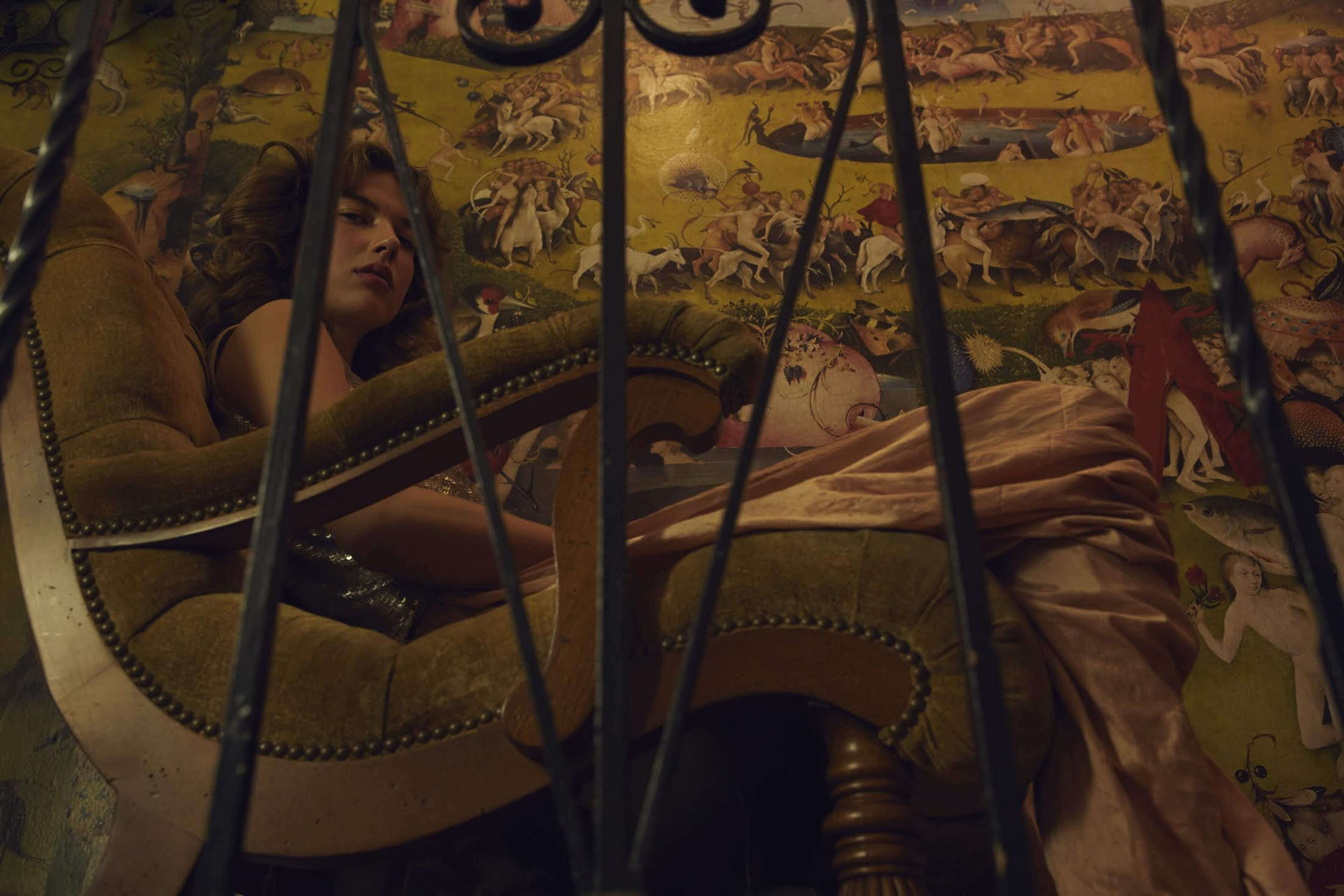 A woman in a low-cut dress sits in a large green armchair in front of a colorful mural depicting various mythical creatures and scenes.