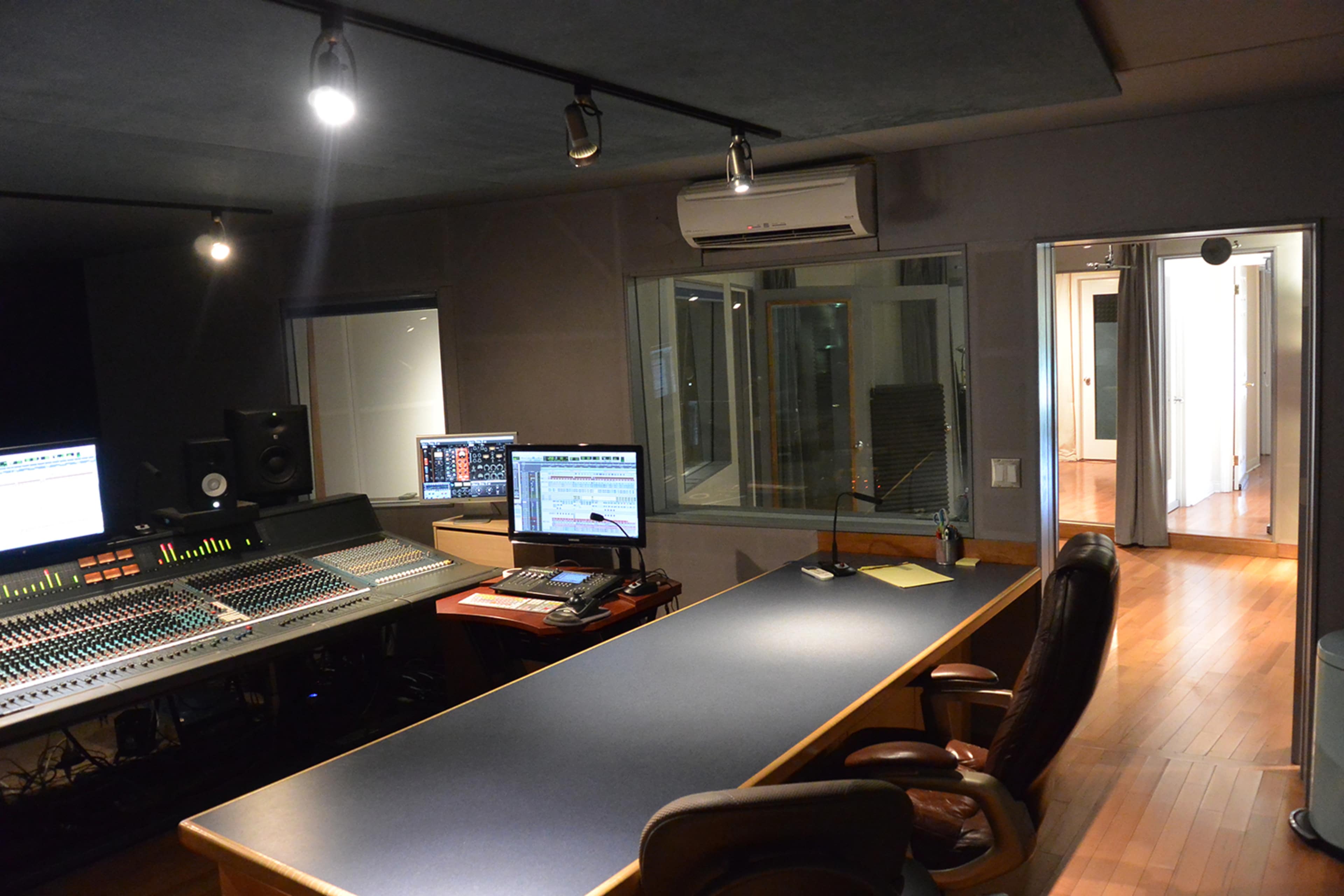 A music recording studio with an audio mixing console, computer monitors, and a large desk, featuring wooden floors and a separate room visible through glass windows.