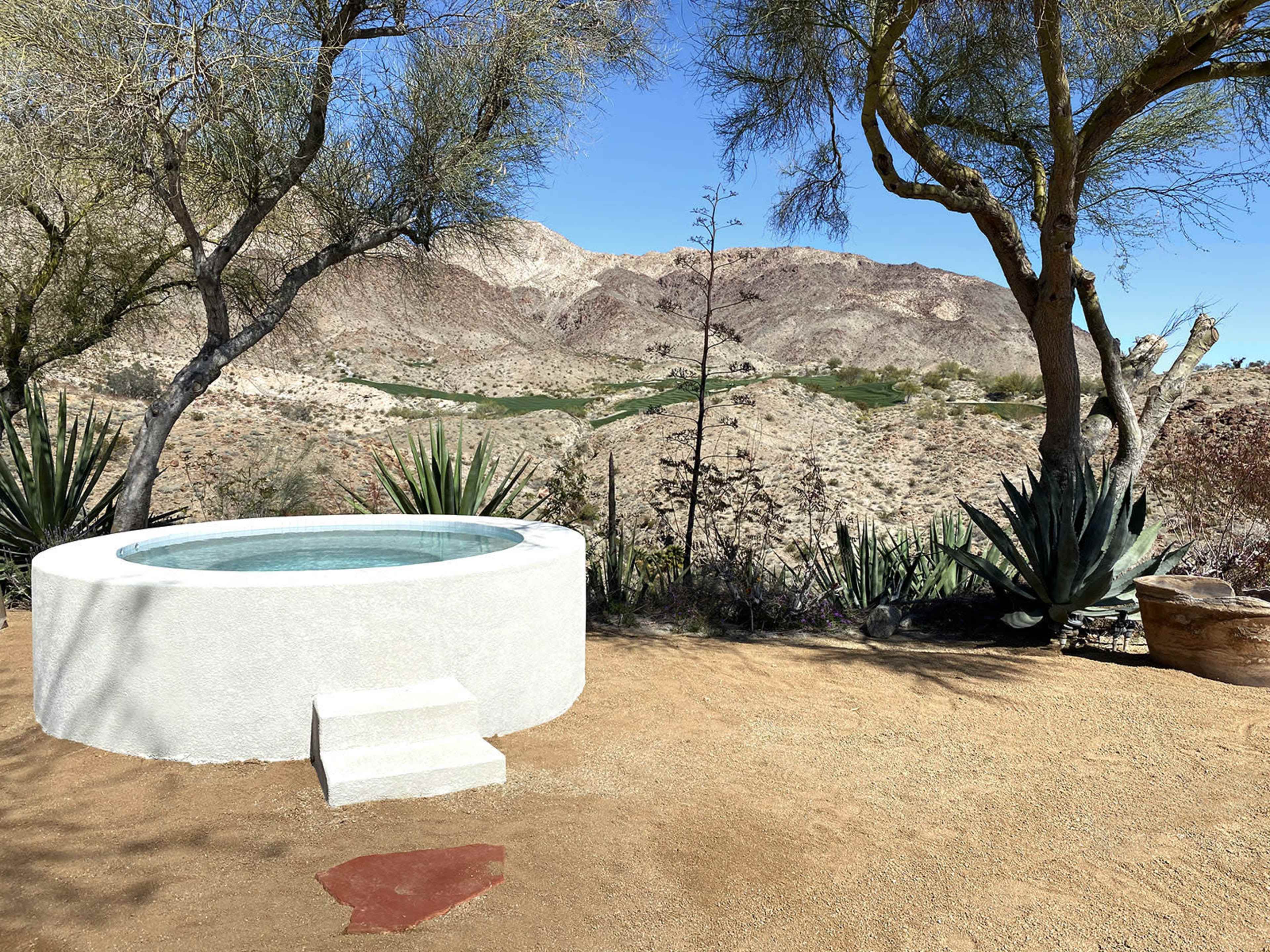 A round hot tub sits atop a sandy area with desert vegetation and mountains in the background.