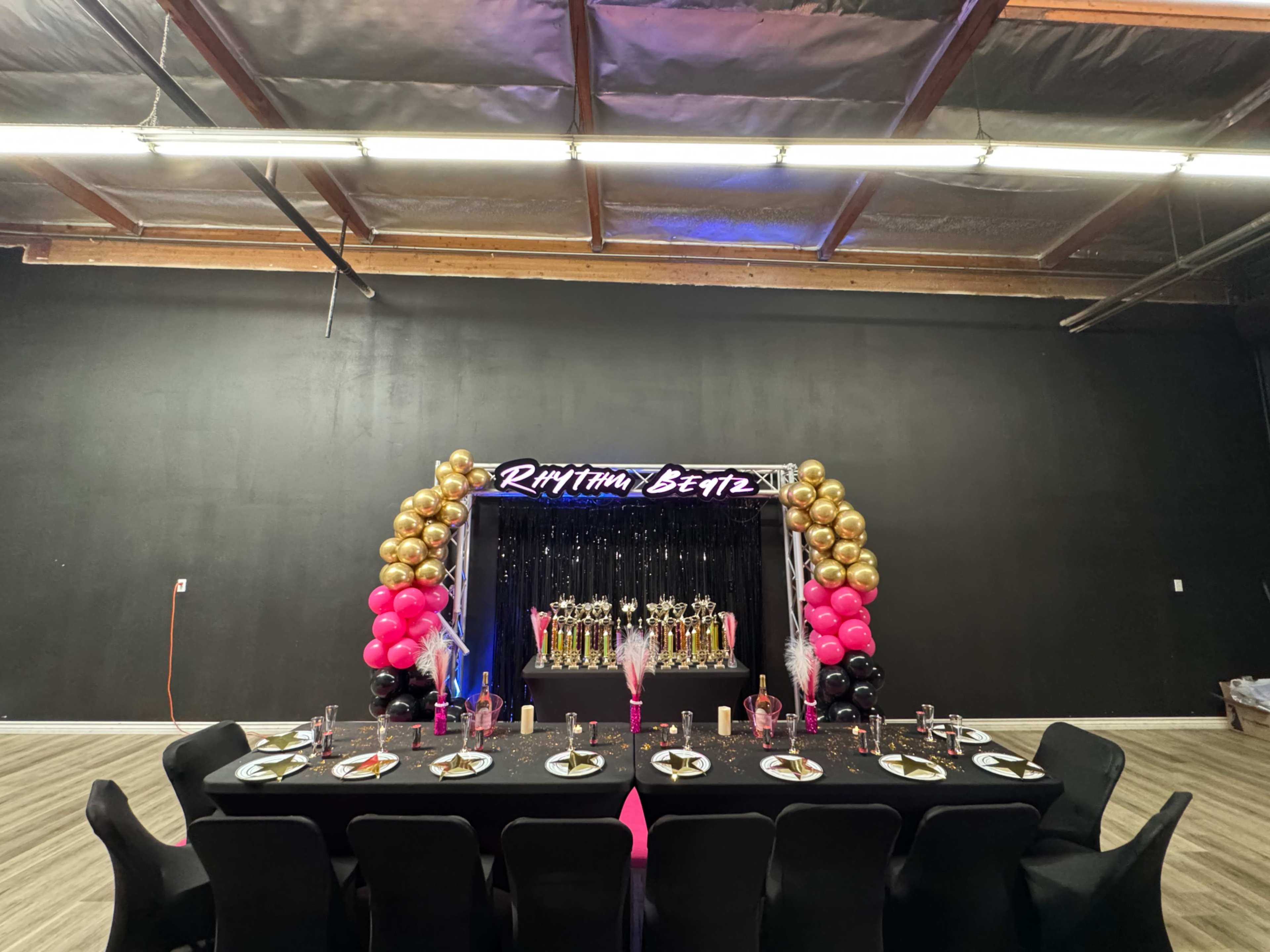 A decorated party space features a table set with black seating and a backdrop of gold and pink balloons with trophies displayed prominently.