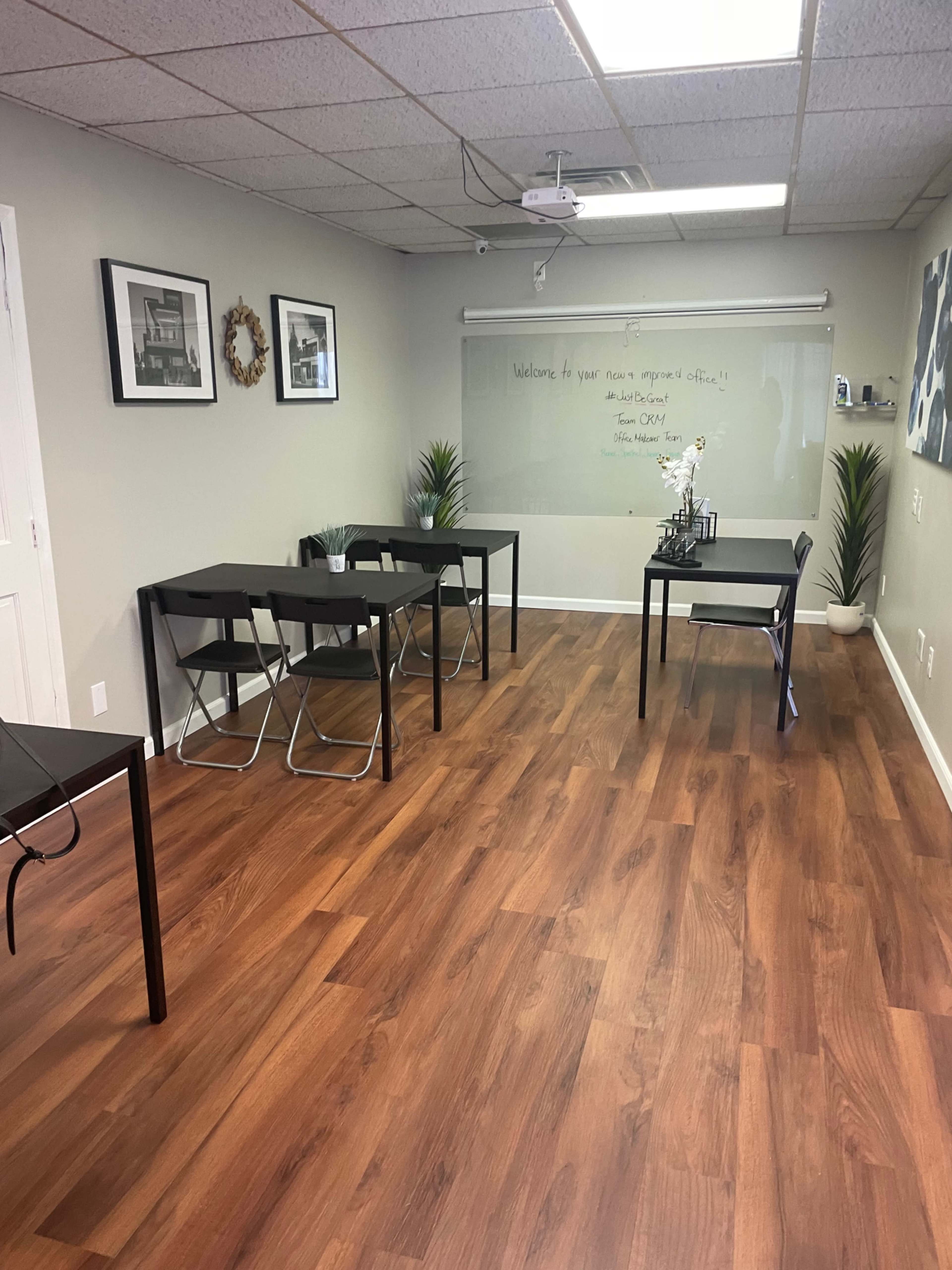 A room with wooden flooring, featuring several black tables and chairs, a whiteboard with a welcome message, and wall decorations.