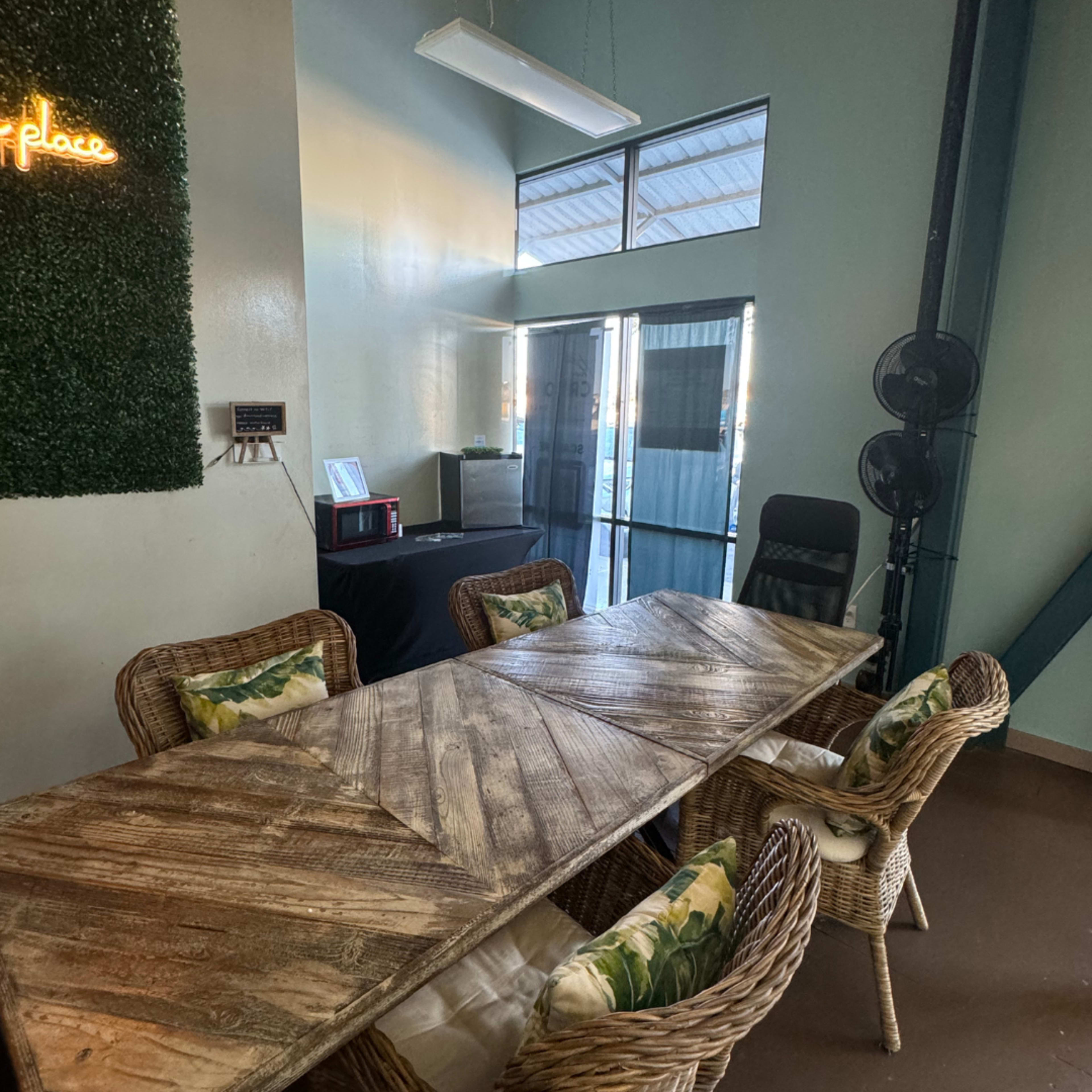 A long wooden table surrounded by wicker chairs is set in a brightly lit room with large windows and a green wall decoration.