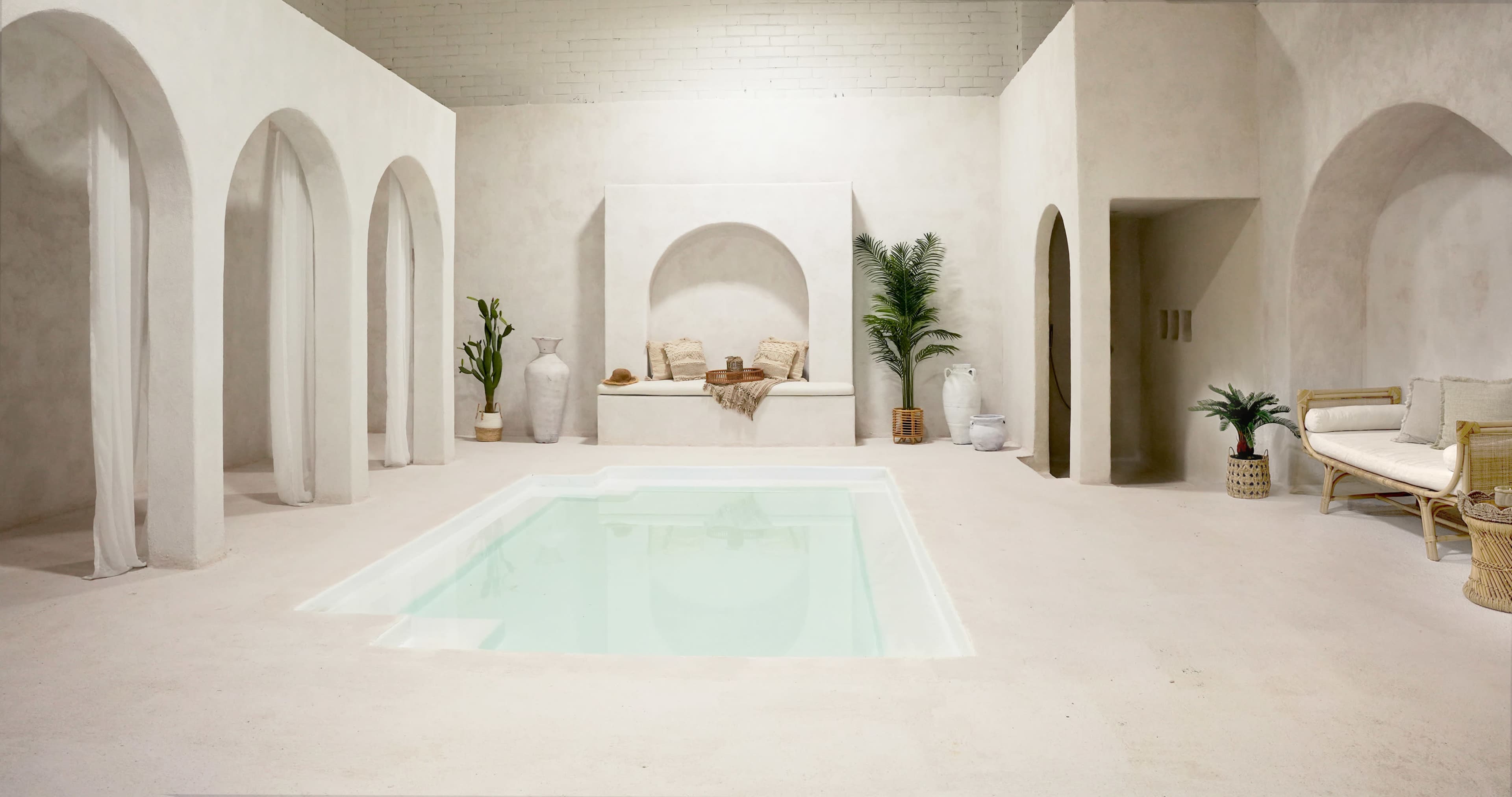 A minimalist interior space featuring a small pool, white walls, and several archways, with potted plants and seating arrangements.