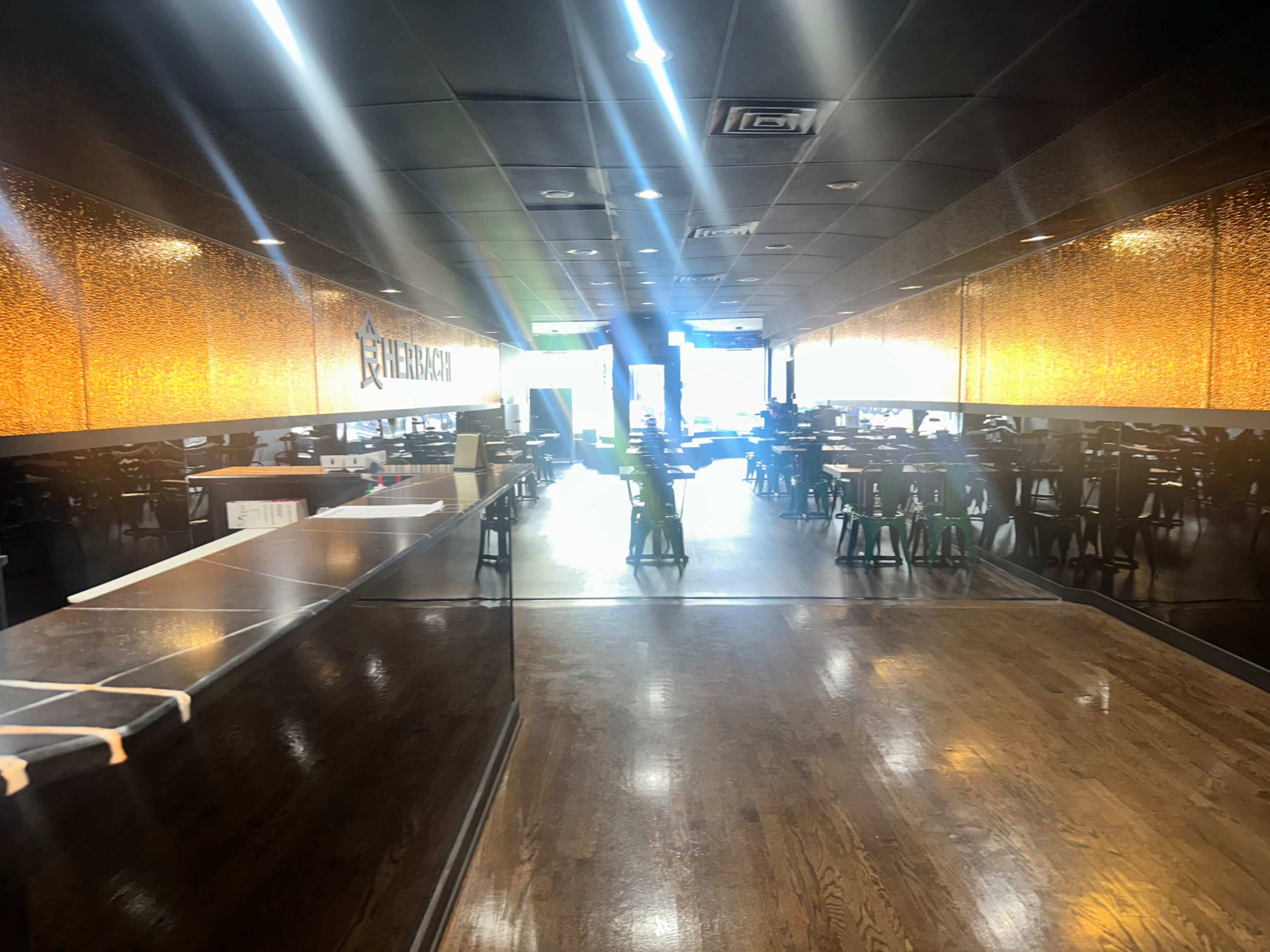 The image shows a bright, spacious restaurant interior with a wooden floor, rows of tables and chairs, and a bar area on one side.