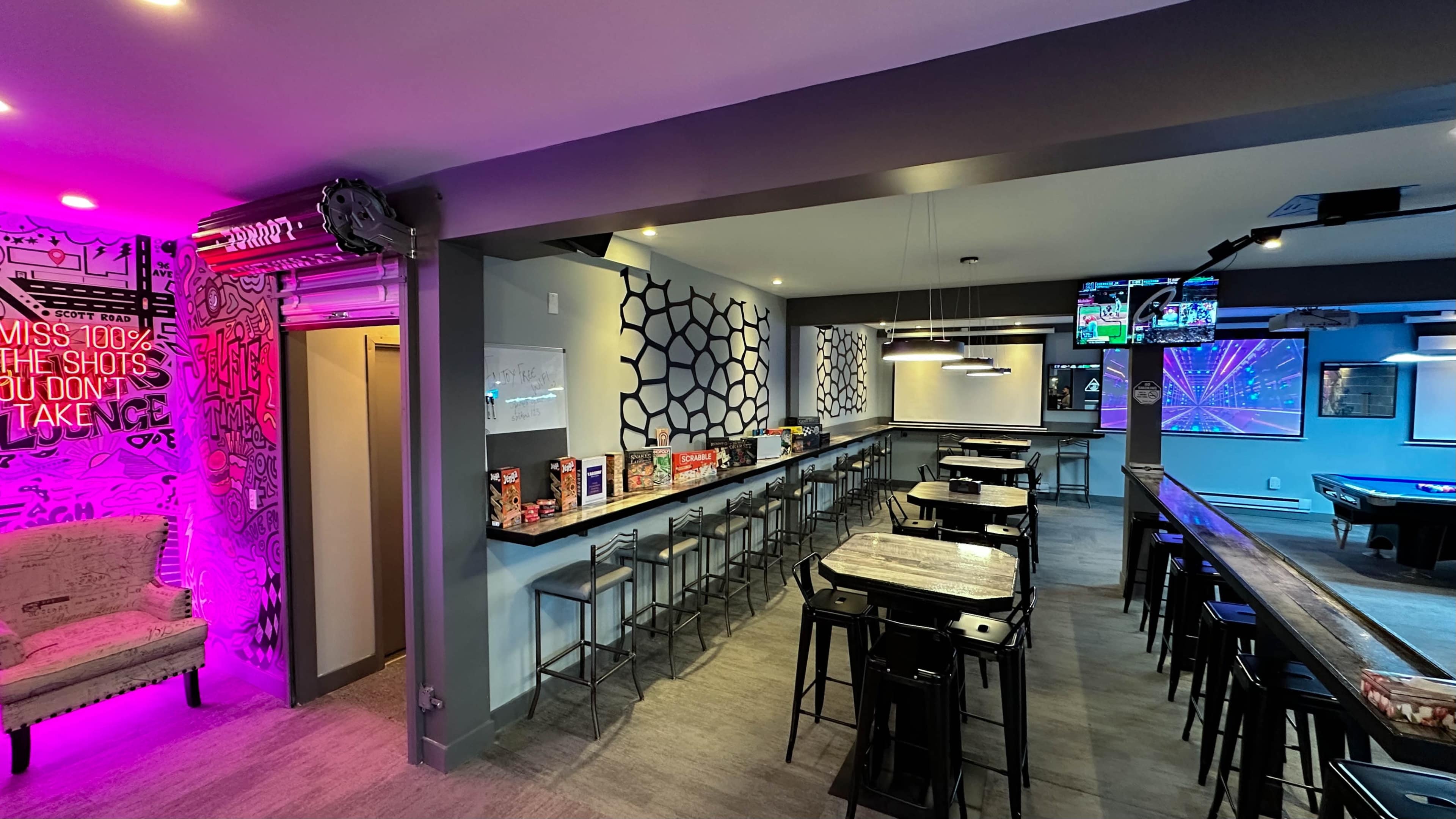A modern recreational space with high-top tables and bar stools, featuring gaming equipment and large screens.