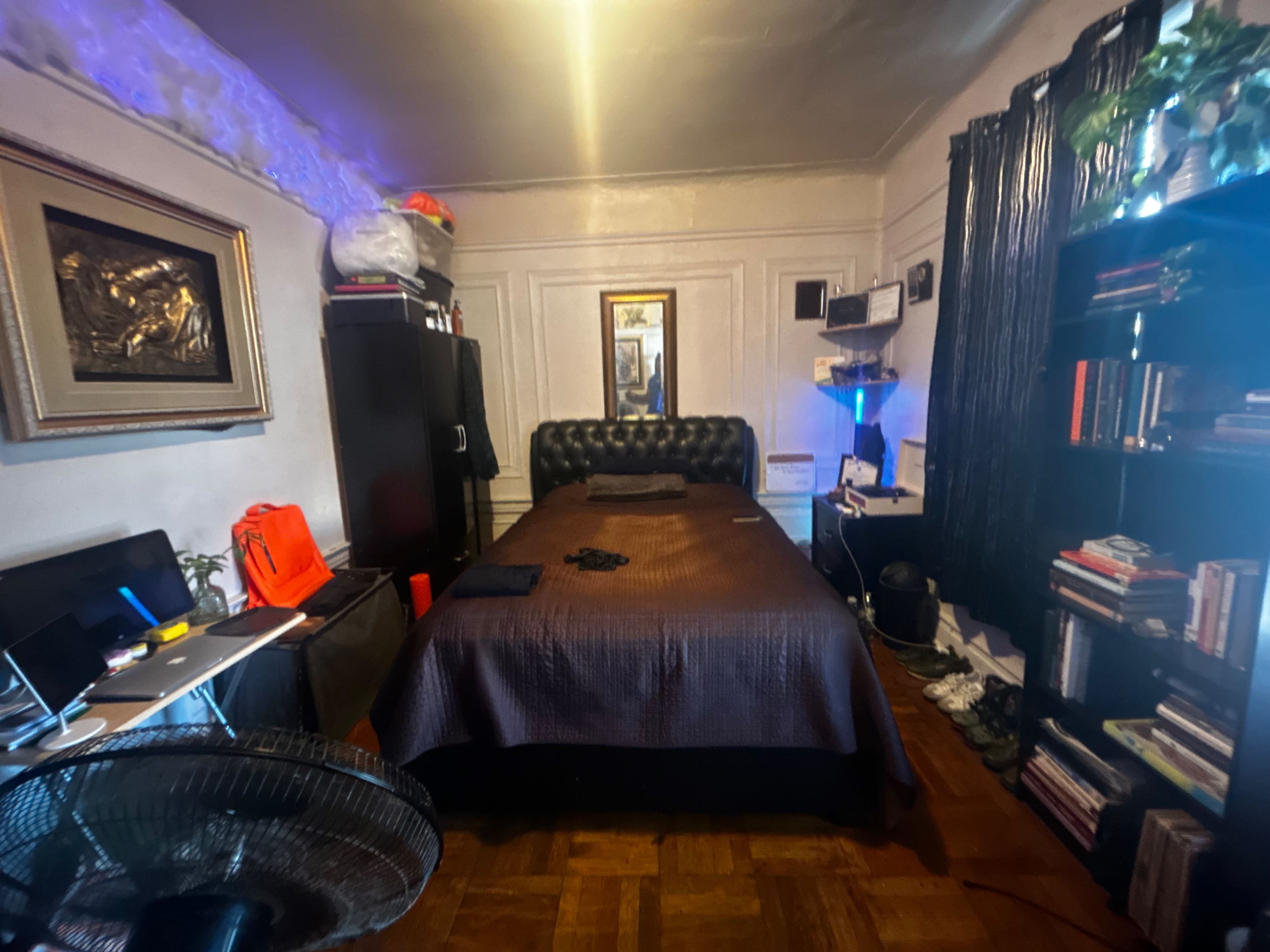 A small bedroom with a black tufted bed, a desk with a laptop, shelves filled with books, and a large mirror on the wall.