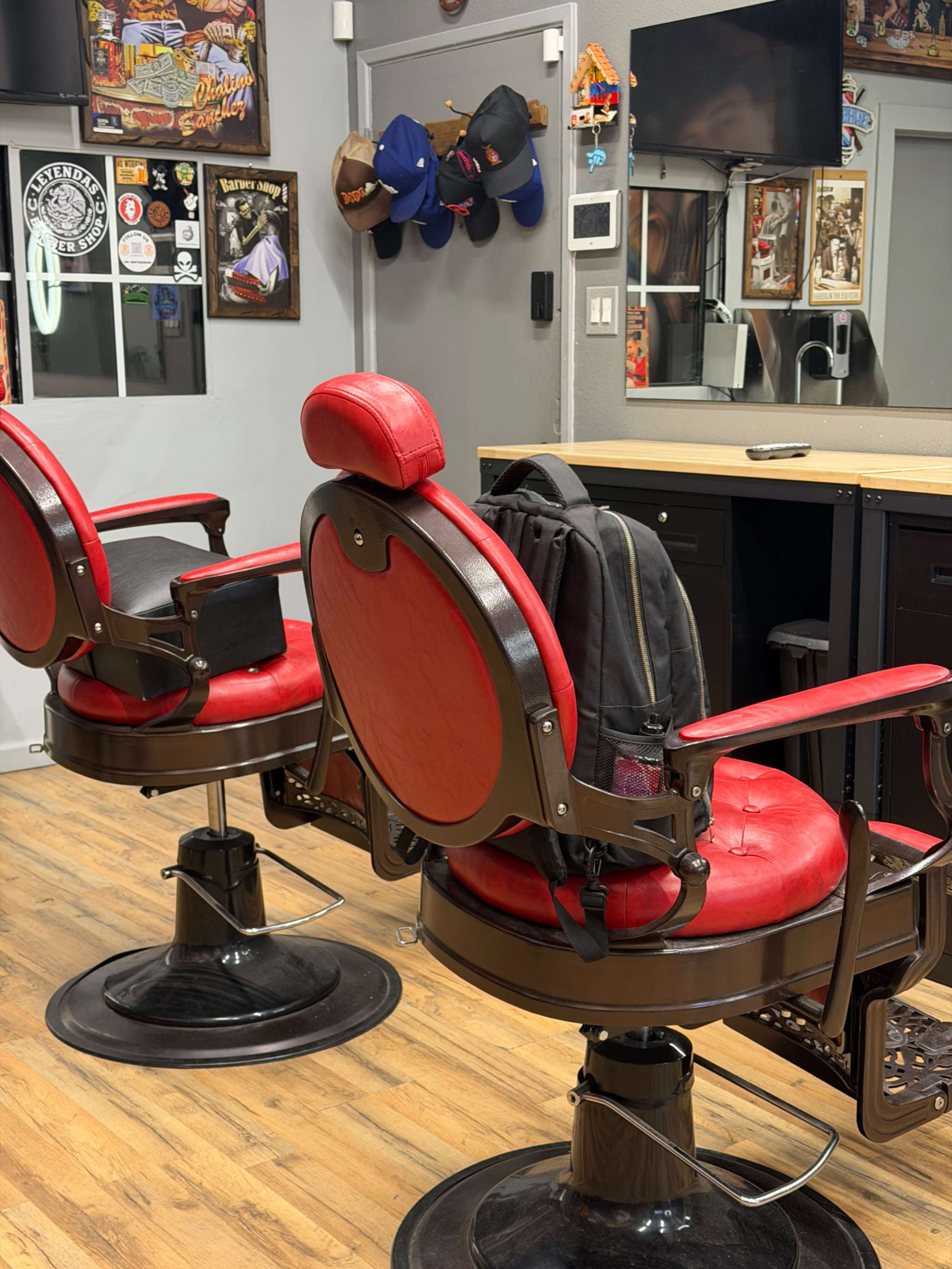 The image shows two red barber chairs in a barbershop, with a backpack placed on one chair and various baseball caps hanging on the wall.