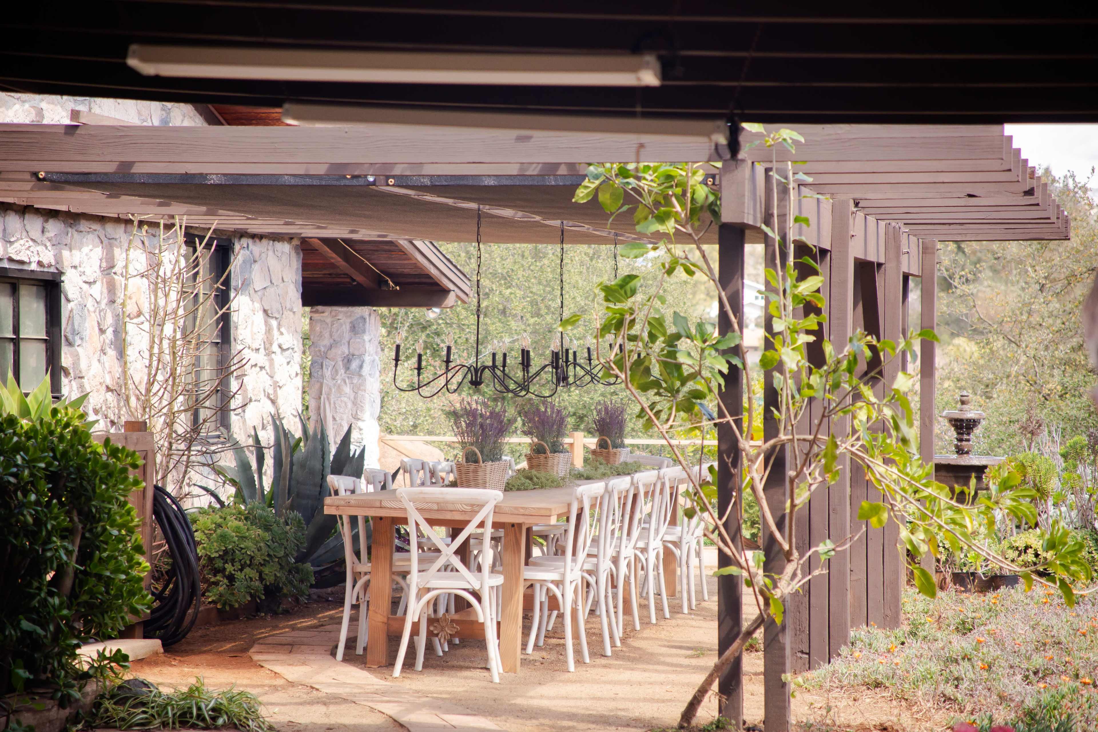 A long wooden dining table with white chairs is set up under a pergola next to a stone building surrounded by greenery.