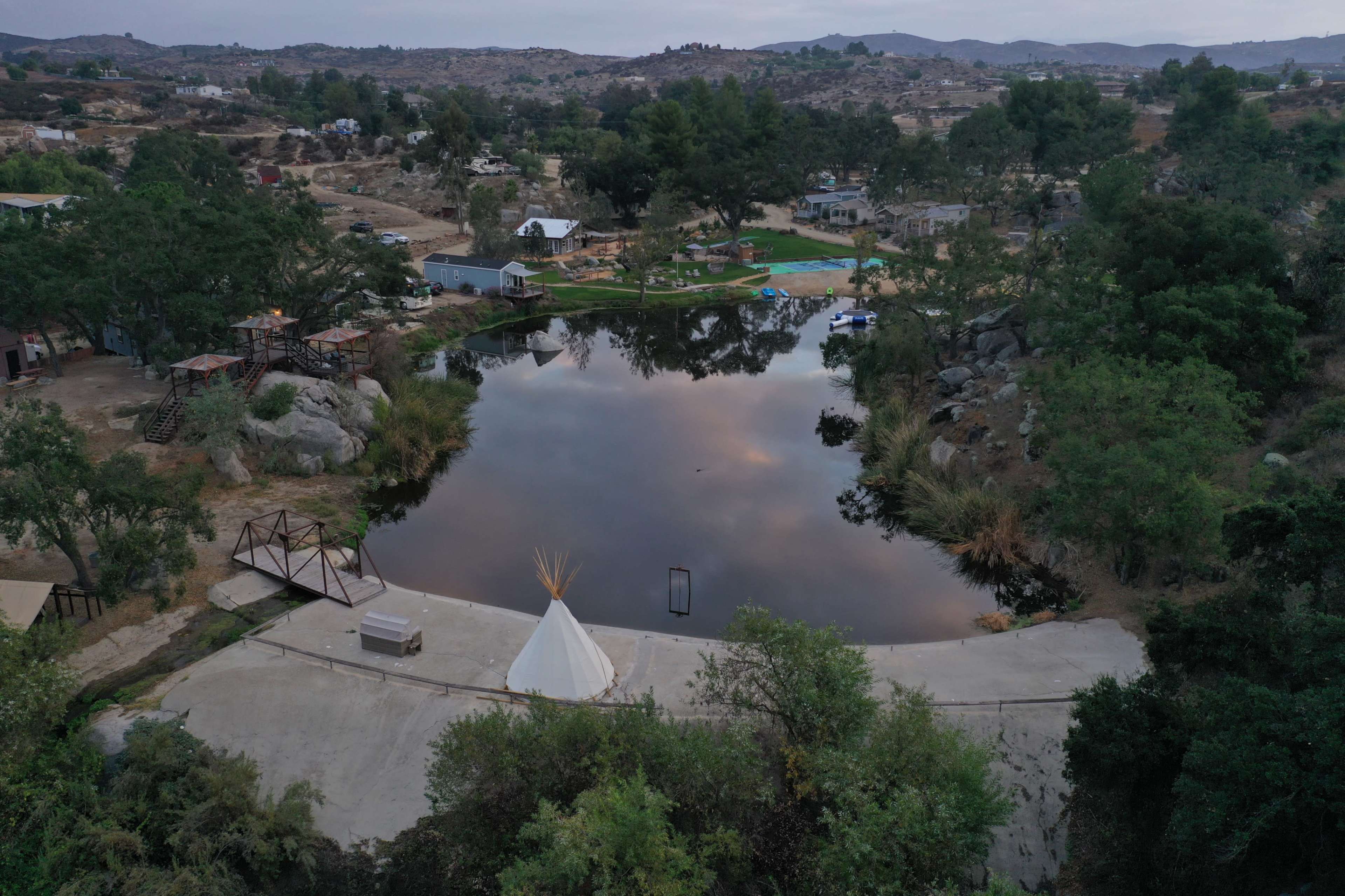 The image shows a tranquil pond surrounded by trees, with a teepee structure and pathways visible along the shore.