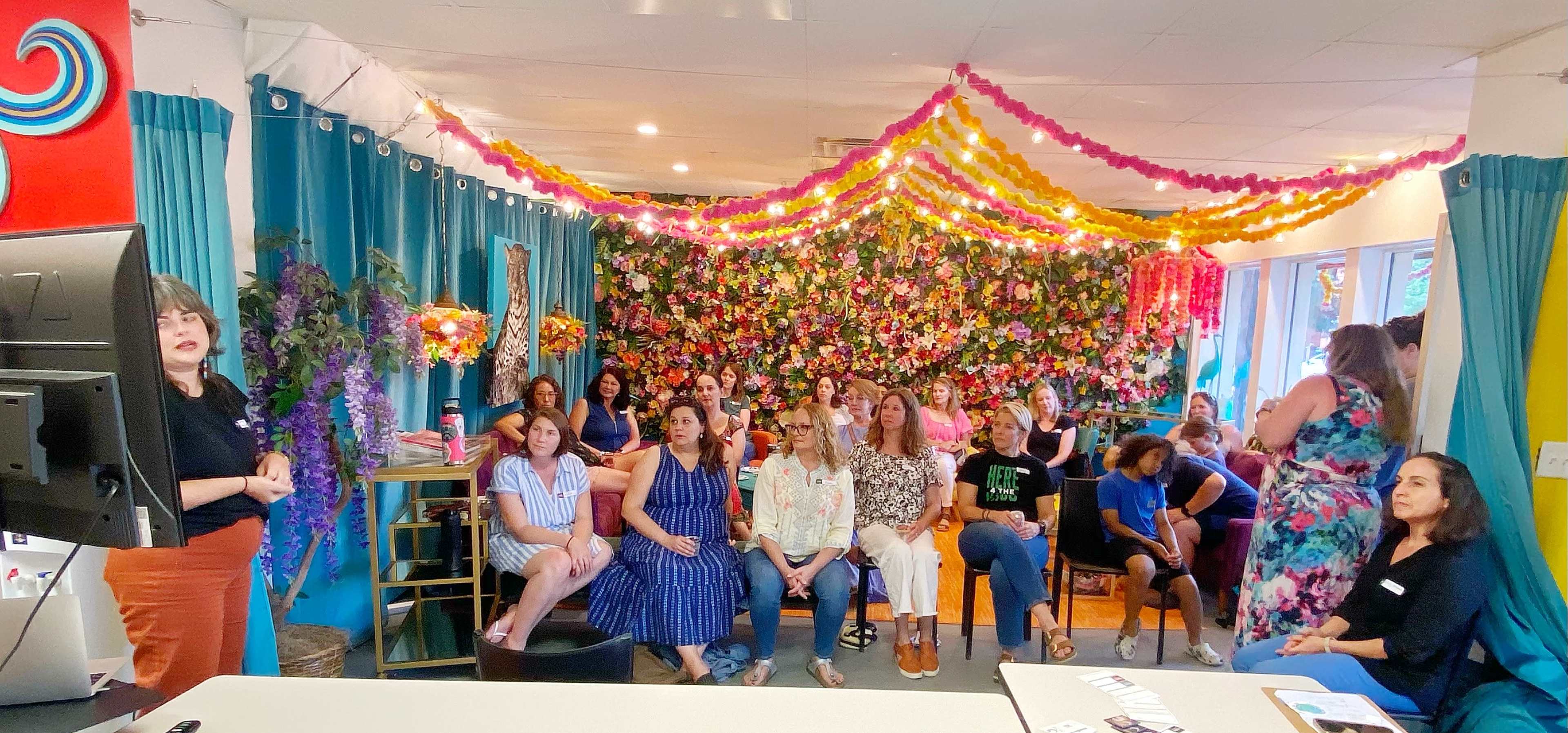 A group of people is seated in a colorful room decorated with a floral backdrop and string lights, listening to a speaker at the front.