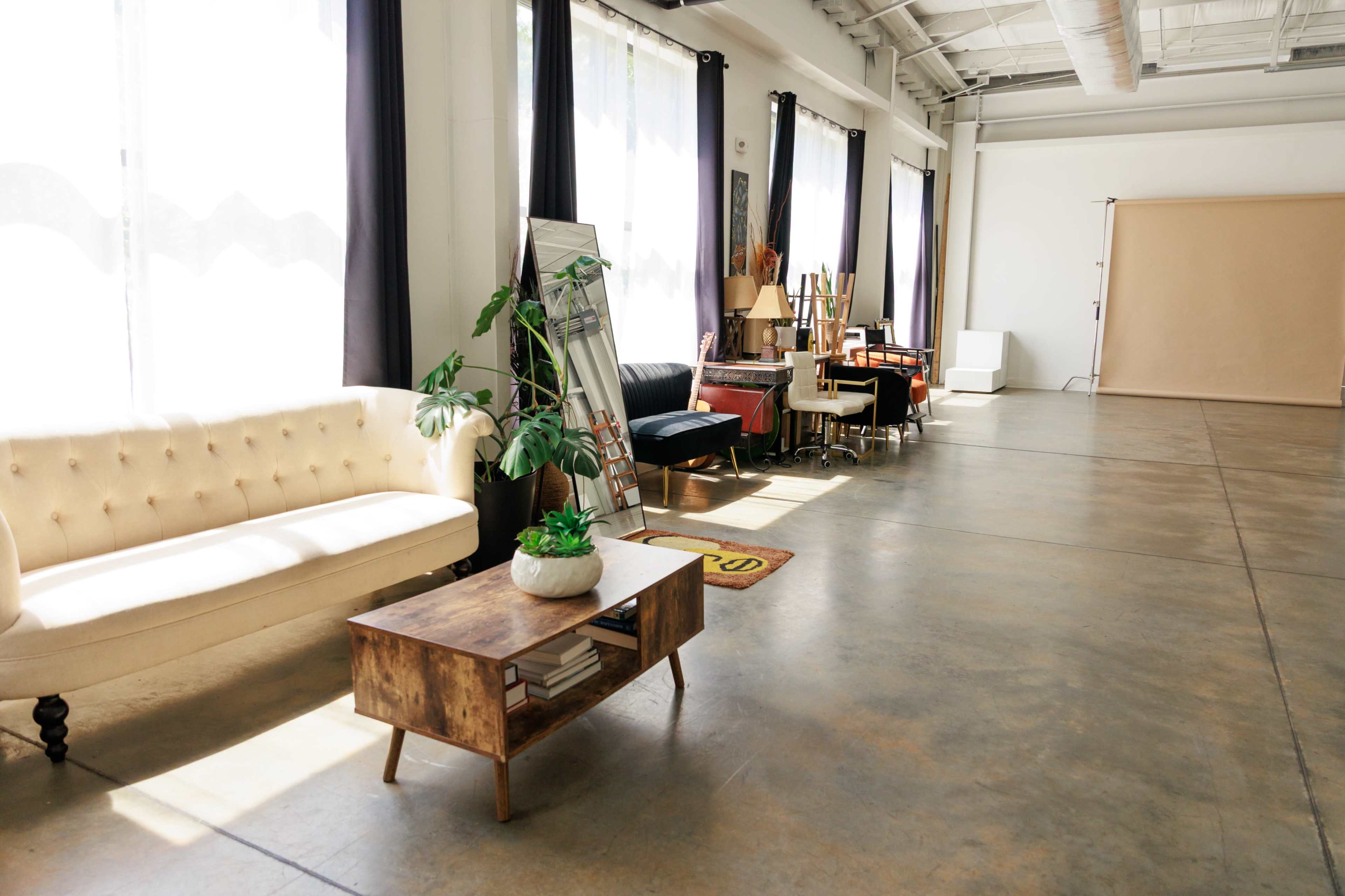 The image shows a spacious, well-lit interior featuring a mix of furniture, including a tufted sofa, a coffee table, and several seating areas, arranged along one side of a concrete floor.