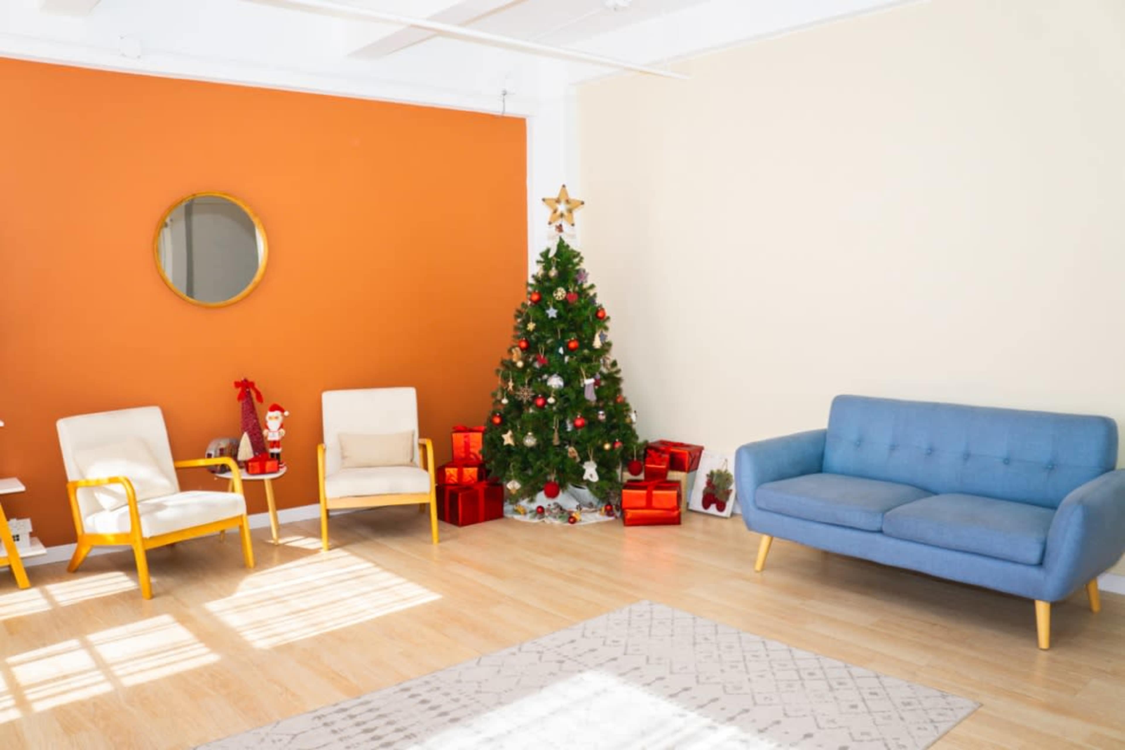 The image shows a cozy room featuring a Christmas tree decorated with lights and ornaments, accompanied by gifts, a blue sofa, and yellow-accented chairs against an orange and cream backdrop.