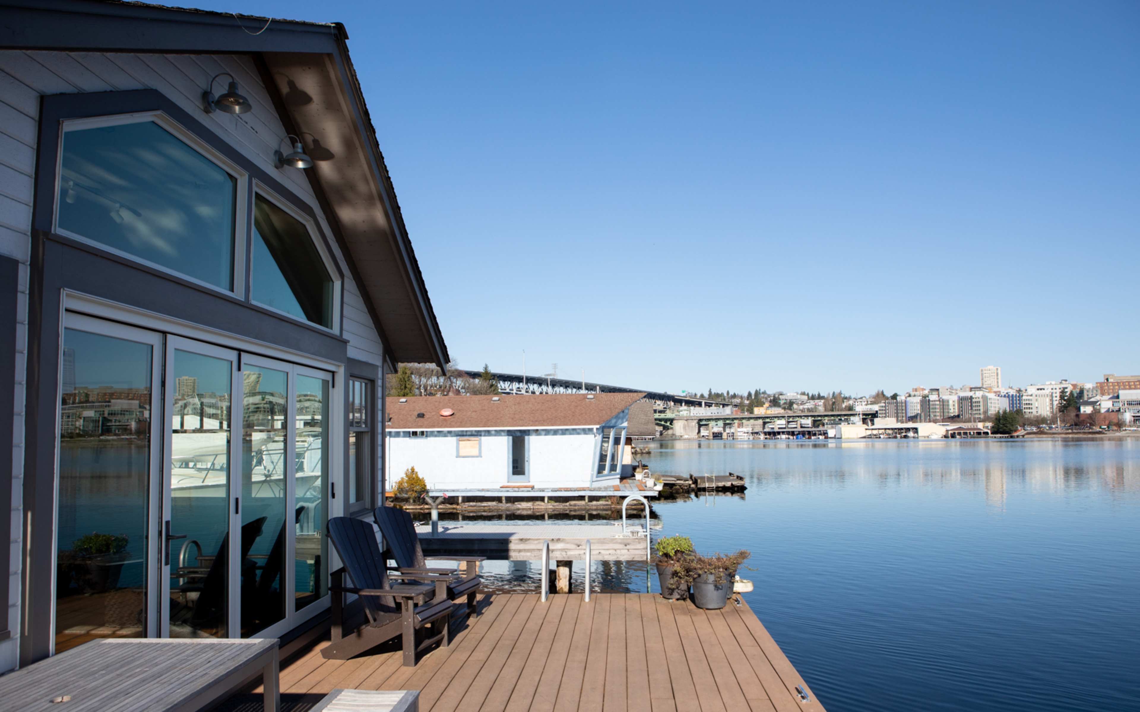 A wooden deck with blue chairs overlooks a calm body of water, reflecting nearby buildings and a clear sky.