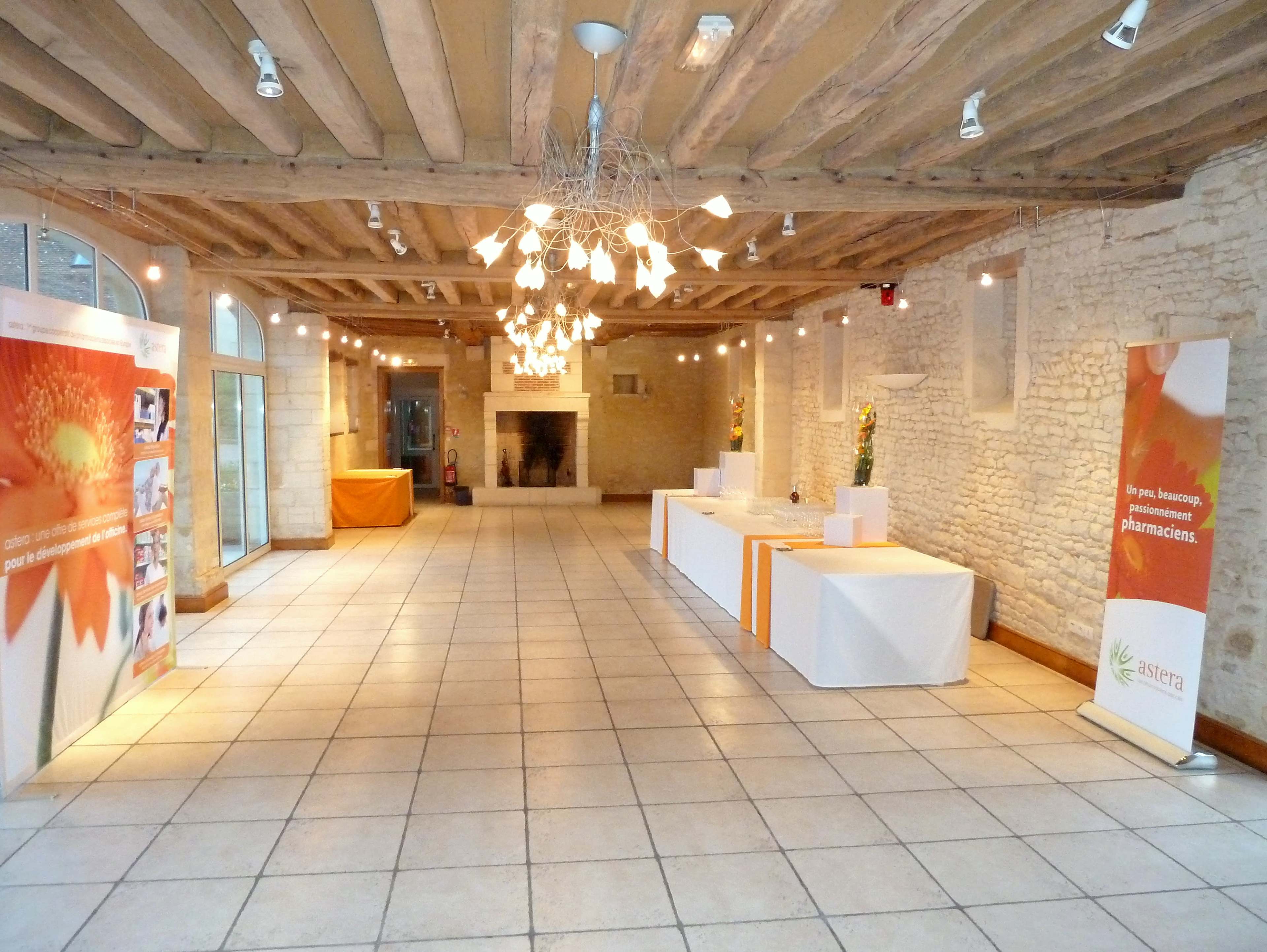 The spacious interior features wooden beams, white stone walls, and a central chandelier, with tables set up along one side for an event.
