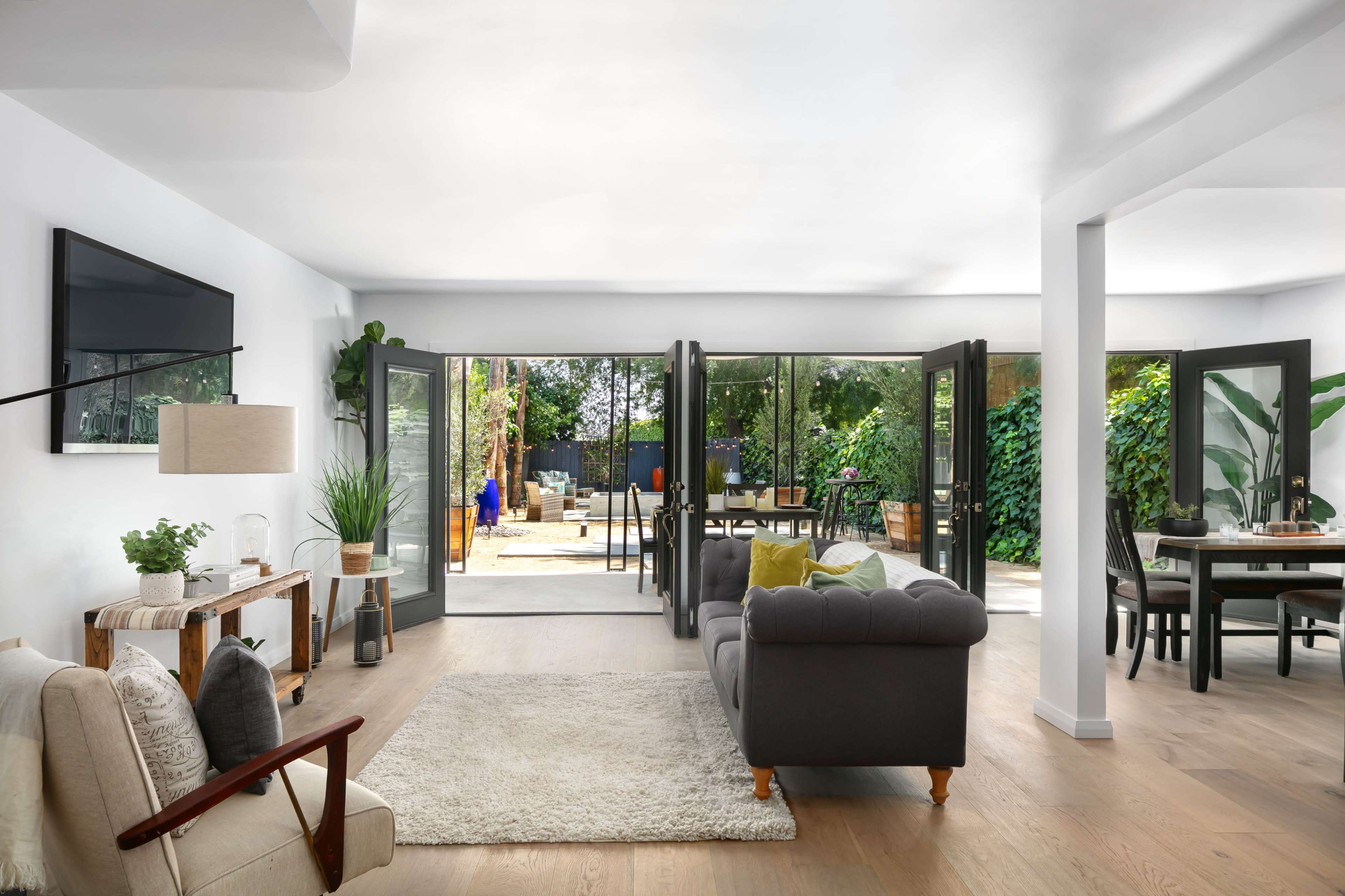 A modern living room opens up to a garden through large glass doors, featuring a sofa, an armchair, and a dining area visible in the background.
