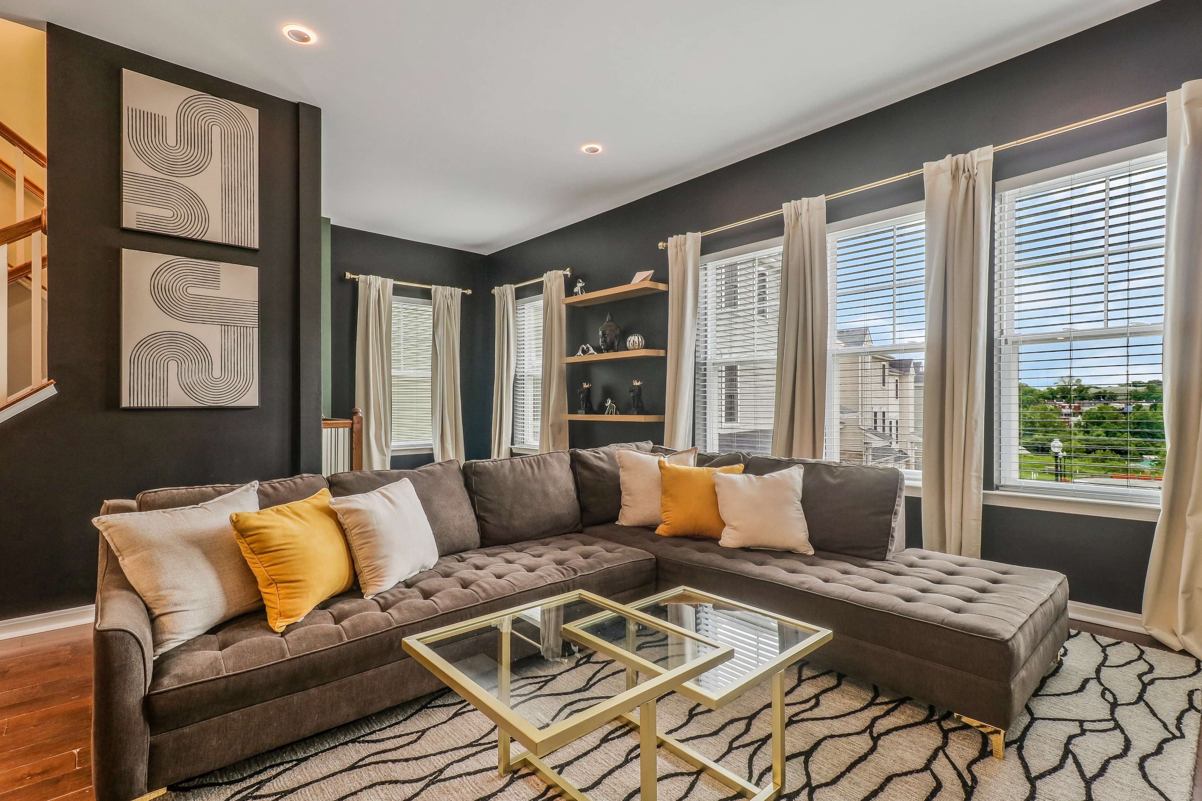 A modern living room with a gray sectional sofa adorned with yellow and white pillows, situated near large windows and decorated with wall art and shelves.