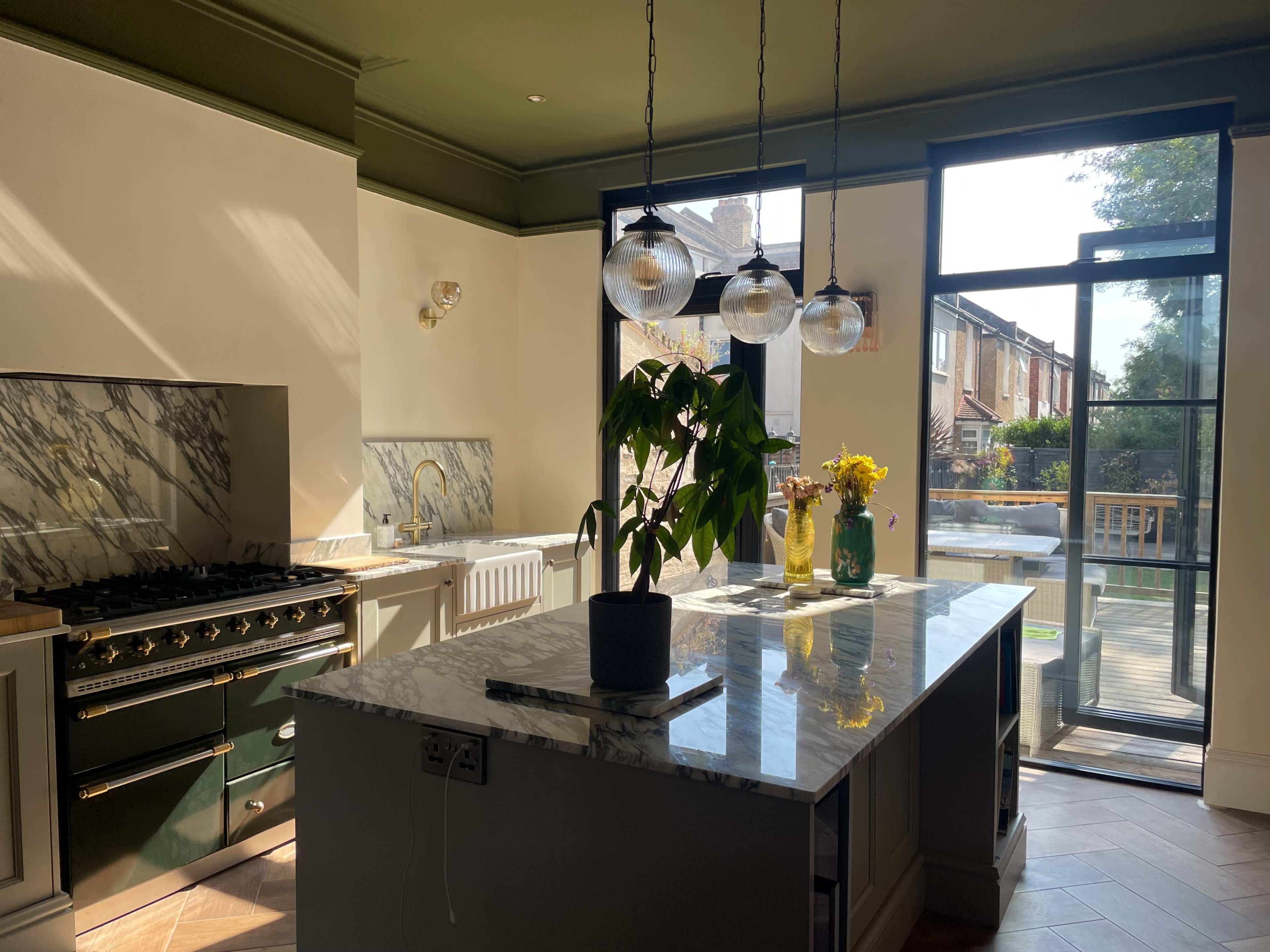 A modern kitchen with a large marble island, a gas stove, and a window letting in natural light, overlooking a garden.