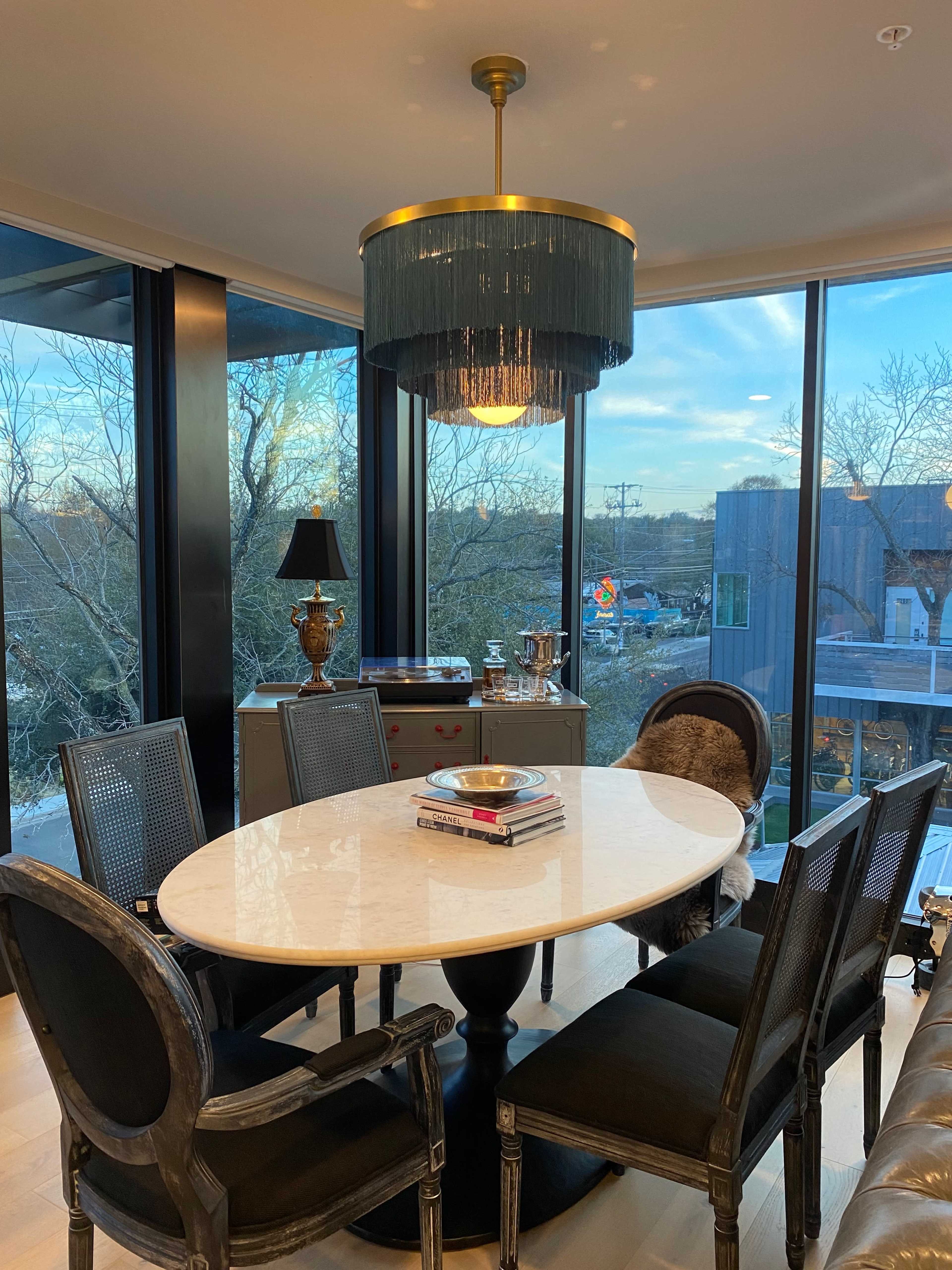 A modern dining area features a round table with a marble top, surrounded by upholstered chairs, under a stylish chandelier, with large windows displaying a view of the outdoors.