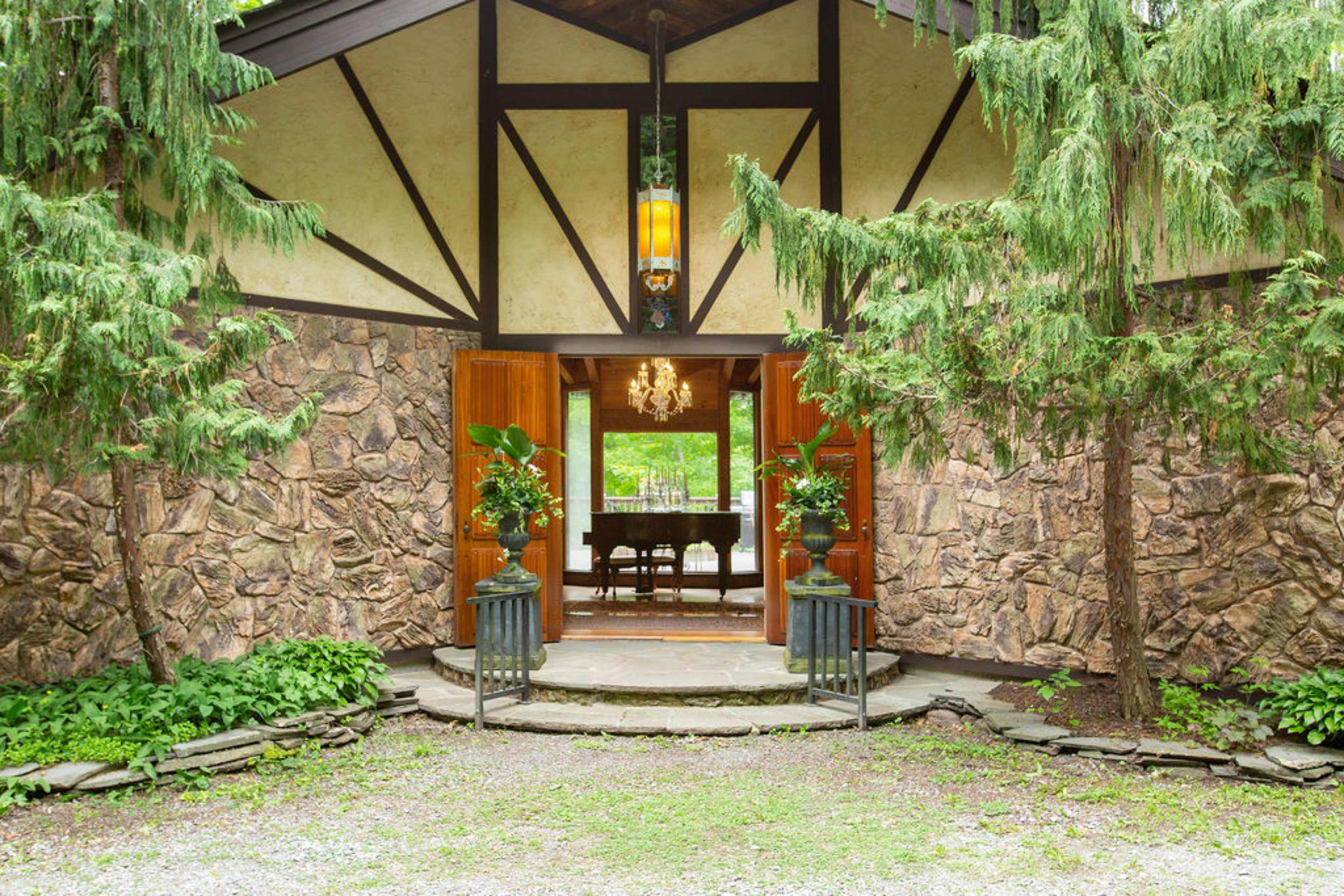 A rustic entrance to a stone and wood cabin, flanked by greenery and featuring a chandelier visible through the open door.